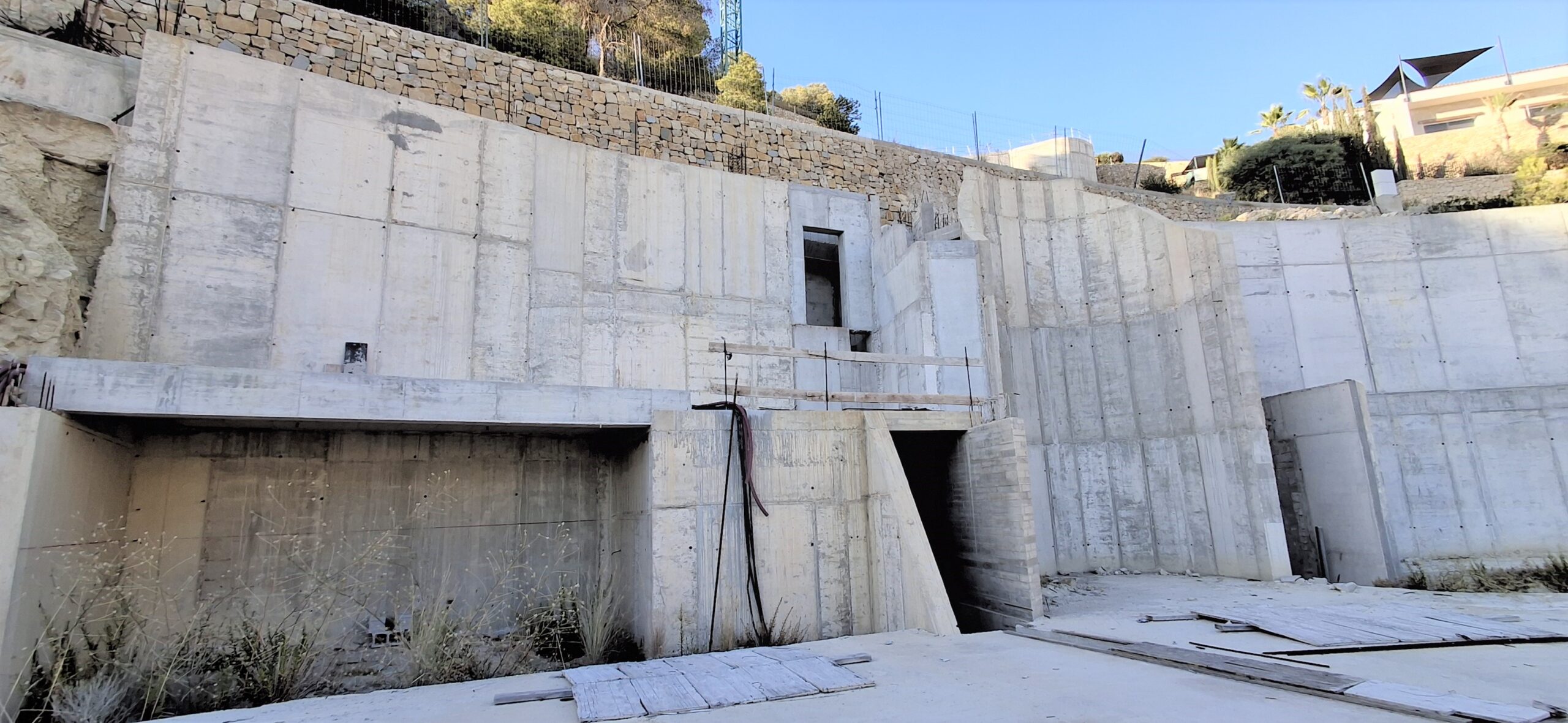 Structural view of the reinforced concrete foundation and retaining walls, featuring integrated stone masonry at the upper level and architectural openings on a sloped site.