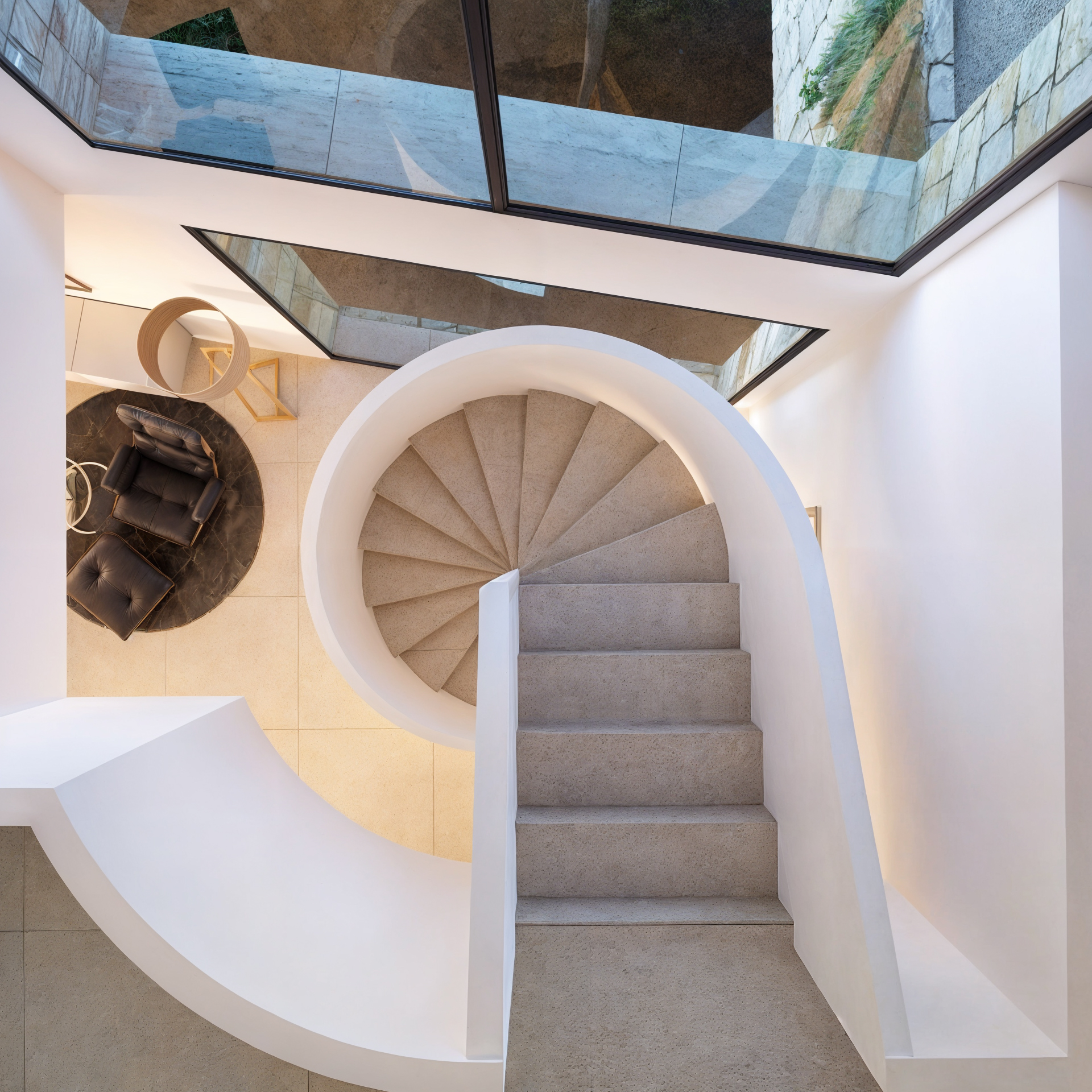 Spiral stone staircase with white curved balustrades viewed from above, featuring large glass windows and a lounge area with a leather armchair below.