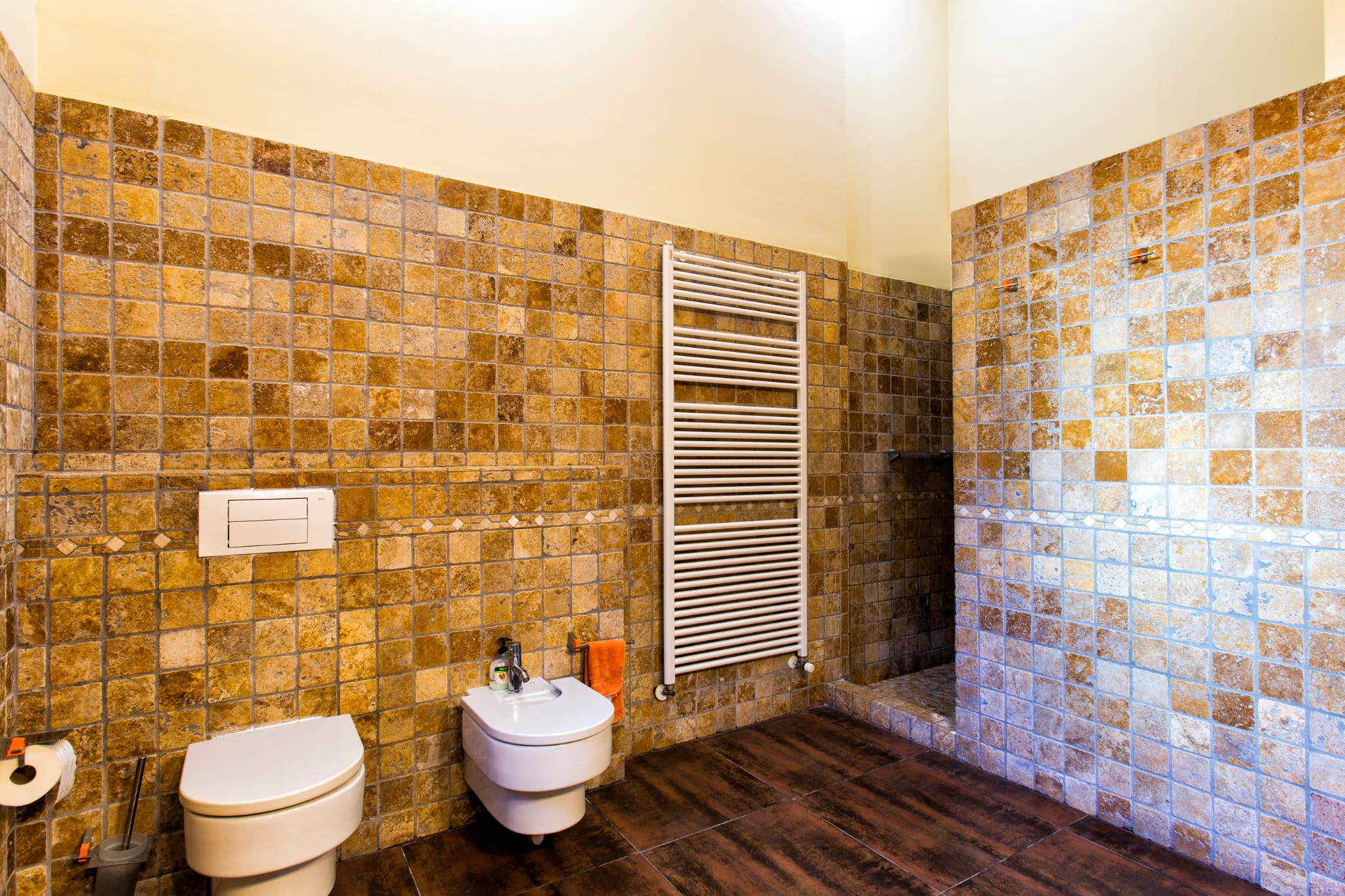 Bathroom featuring earth-toned stone wall tiles, a wall-mounted toilet and bidet, a white heated towel rail, and dark wood-effect flooring.