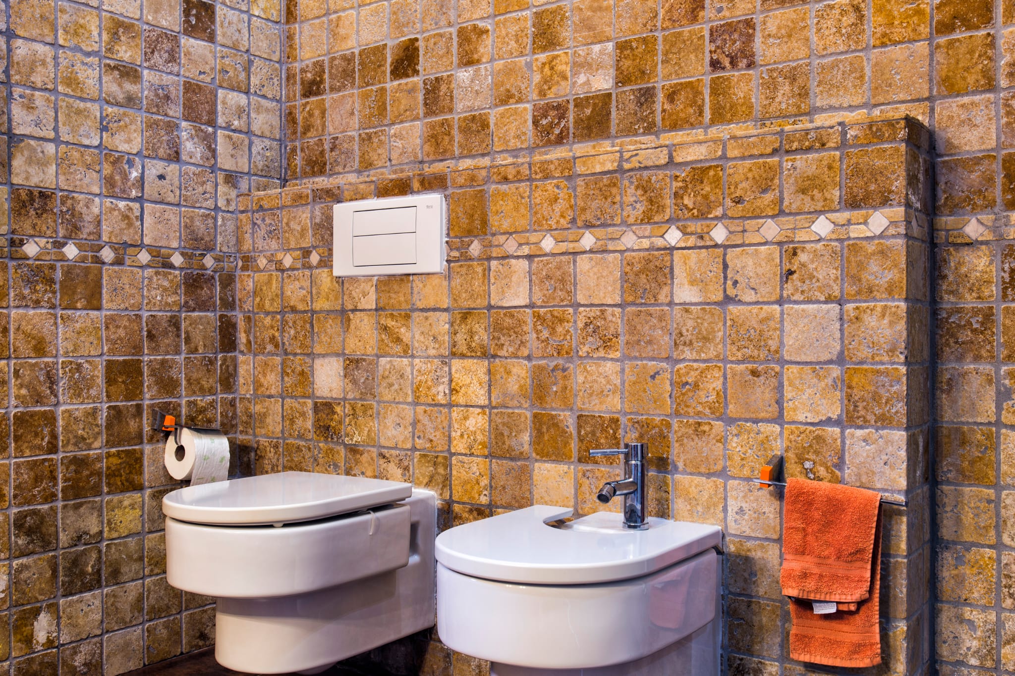 Bathroom featuring rustic earth-toned stone tiles, wall-mounted ceramic toilet and bidet, and a decorative mosaic border.