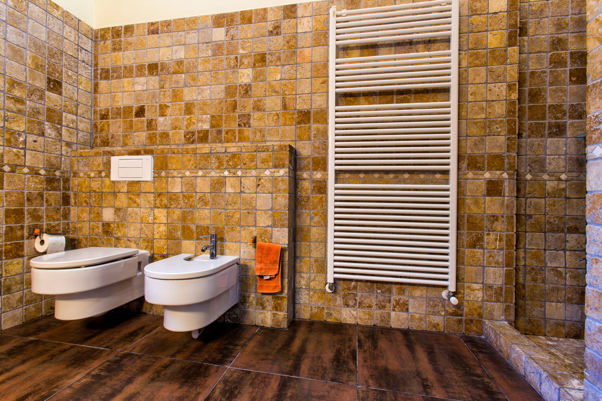 Bathroom featuring wall-mounted toilet and bidet, textured earth-tone mosaic tiles, and a large white heated towel rail against a dark tiled floor.