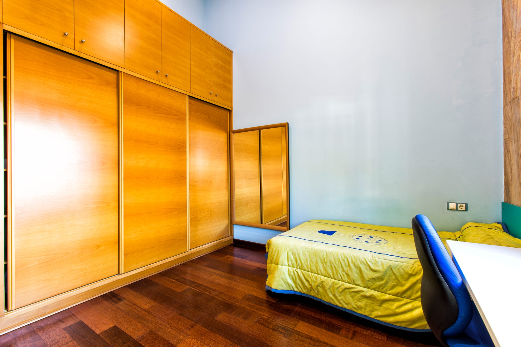 Bedroom featuring a large built-in wooden wardrobe with sliding doors, dark parquet flooring, and a single bed next to a workspace area.
