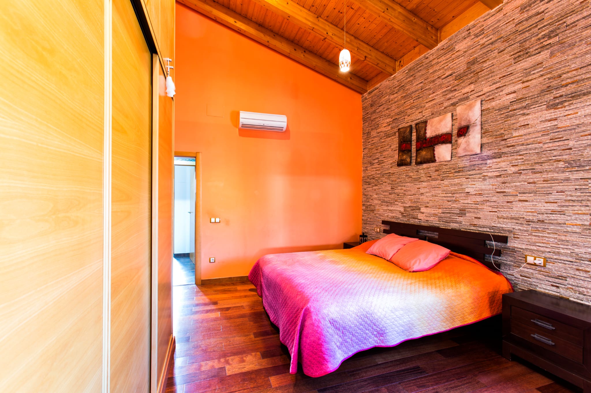Bedroom featuring a slanted wooden beam ceiling, a textured stone accent wall, and built-in wardrobes. The space includes dark wood furniture and polished parquet flooring.