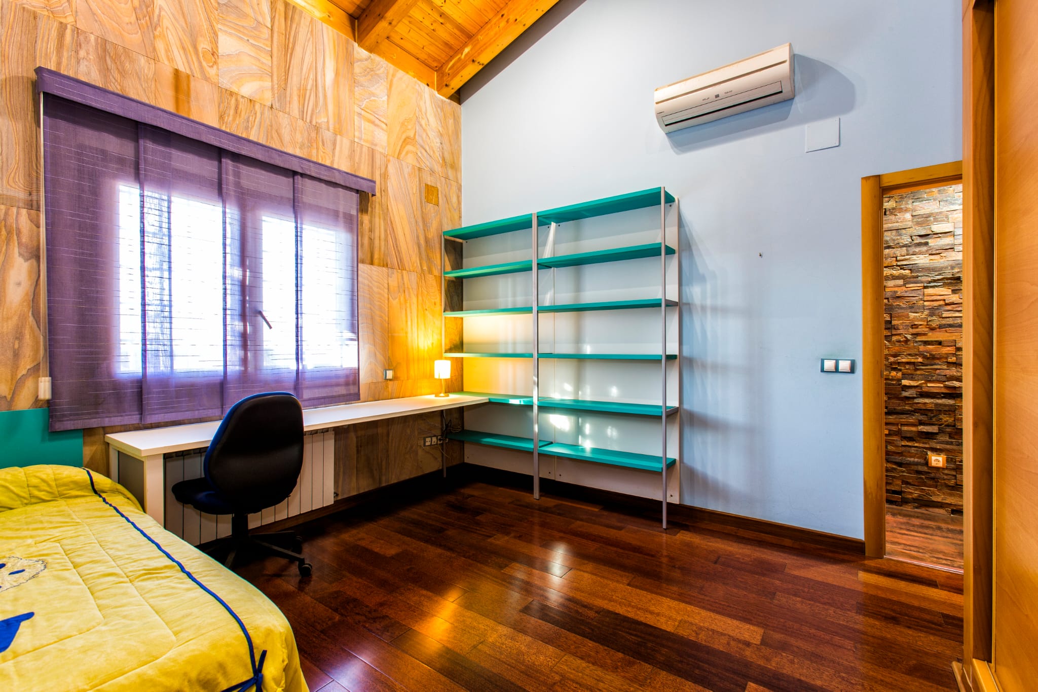Bedroom featuring a study area with a turquoise shelving unit, sloped wooden ceilings, and a white desk under a window with purple blinds.