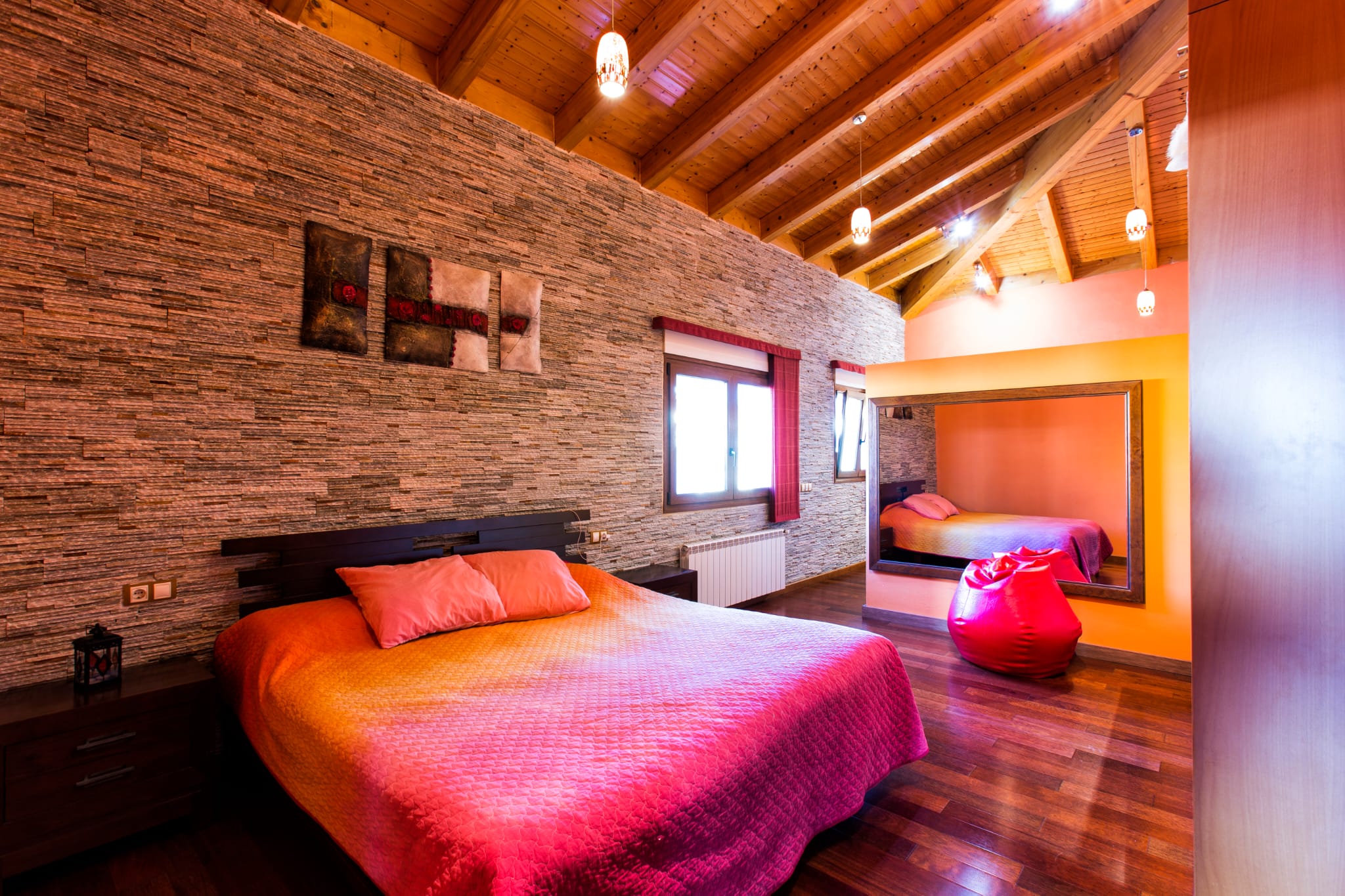 Bedroom featuring a textured stone accent wall, exposed wooden beam ceilings, and dark parquet flooring. Includes a large wall mirror and integrated pendant lighting.