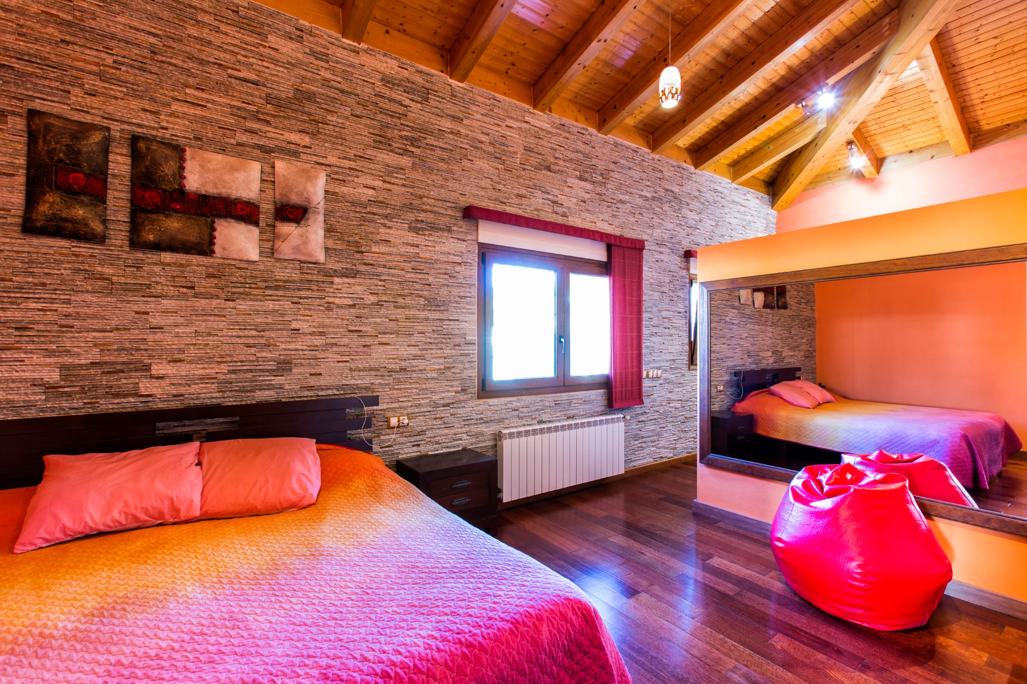 Bedroom featuring a textured stone accent wall, vaulted wooden beam ceilings, and dark hardwood floors. A large wall mirror reflects the bed with vibrant pink bedding.