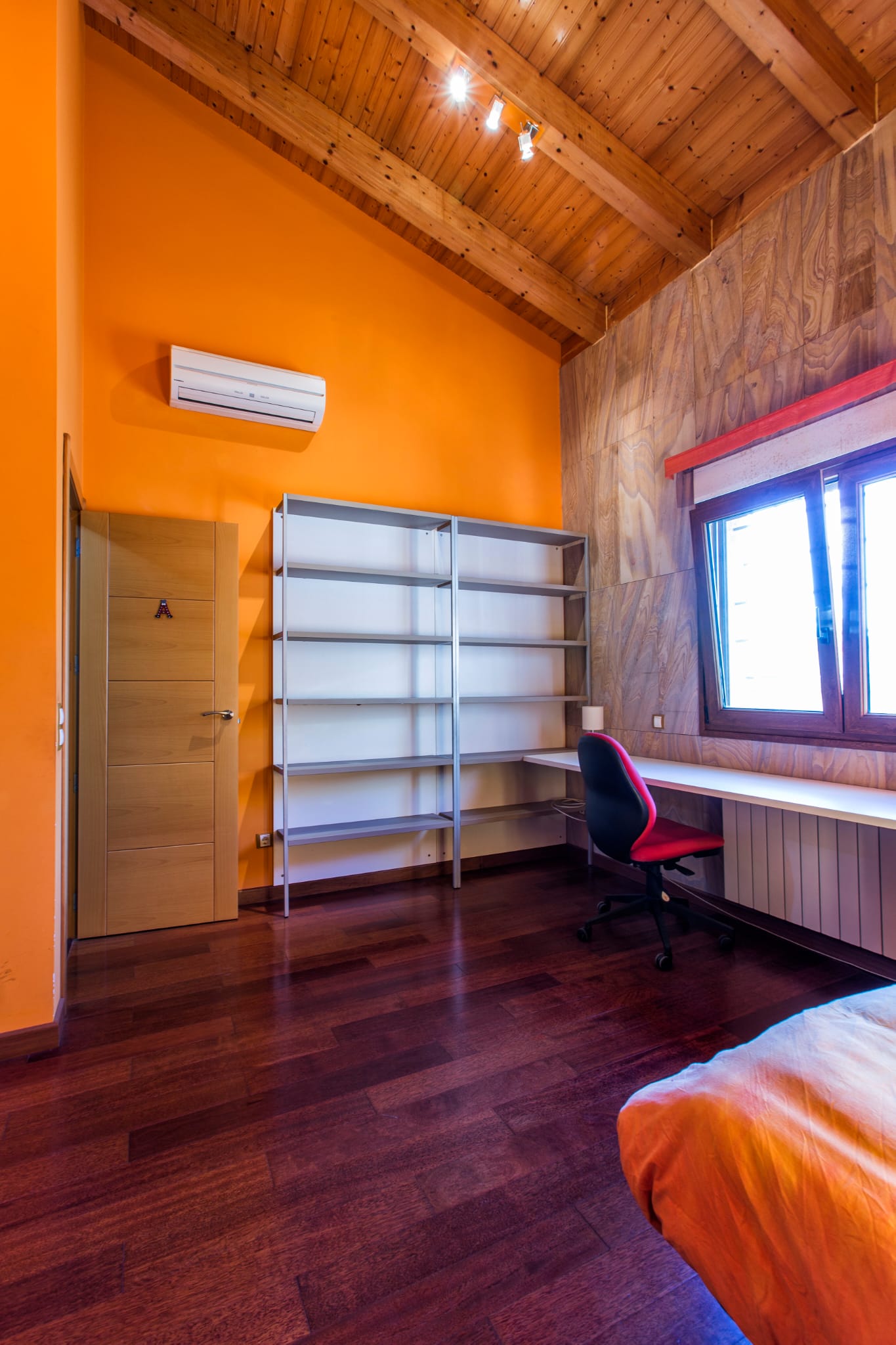 Bedroom featuring vaulted wooden ceilings, orange walls, an integrated study area with a long desk, metal shelving, and dark hardwood flooring.
