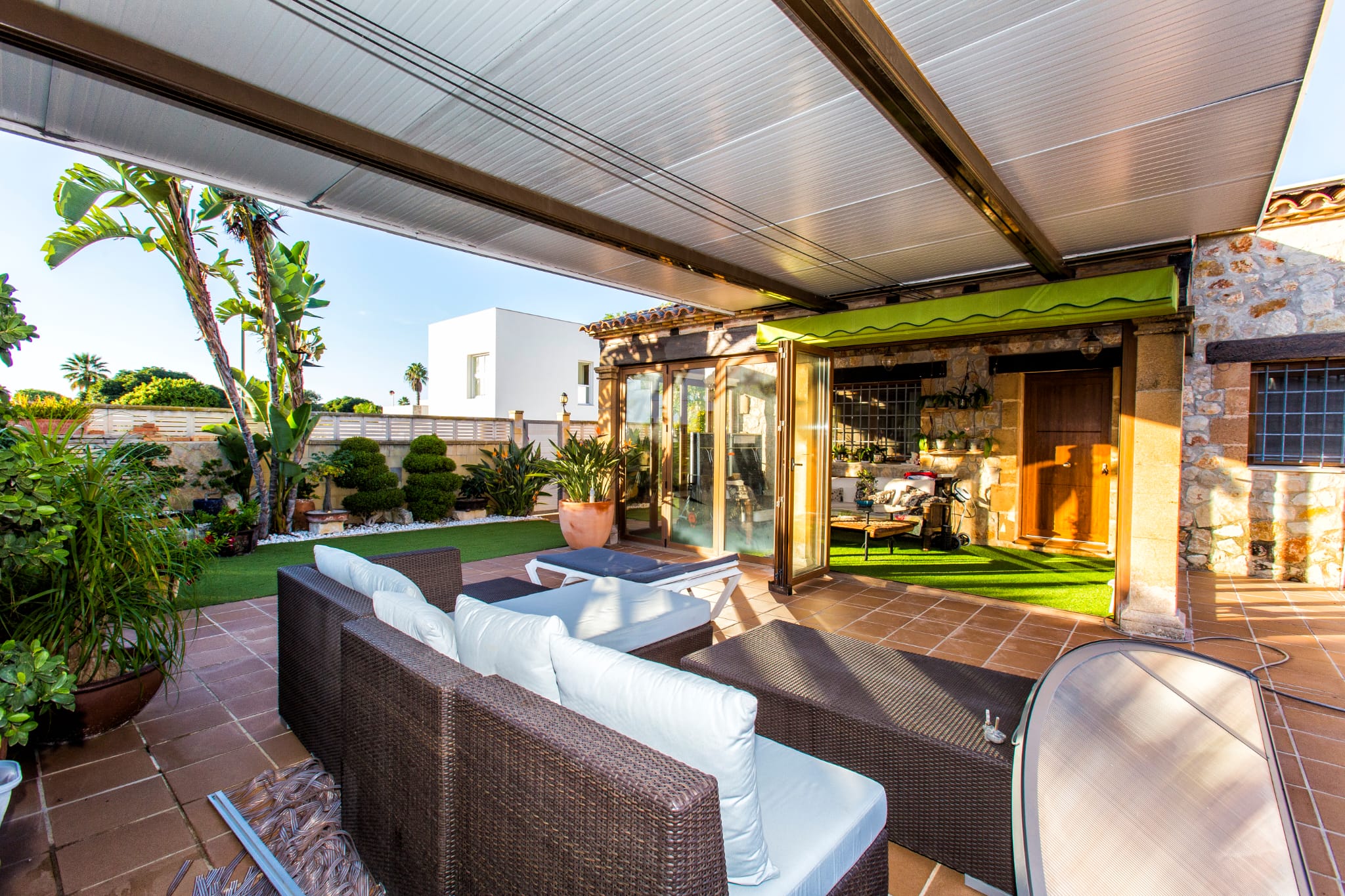 Covered outdoor terrace featuring wicker lounge furniture, terracotta tiling, and a landscaped garden with artificial turf and tropical plants.