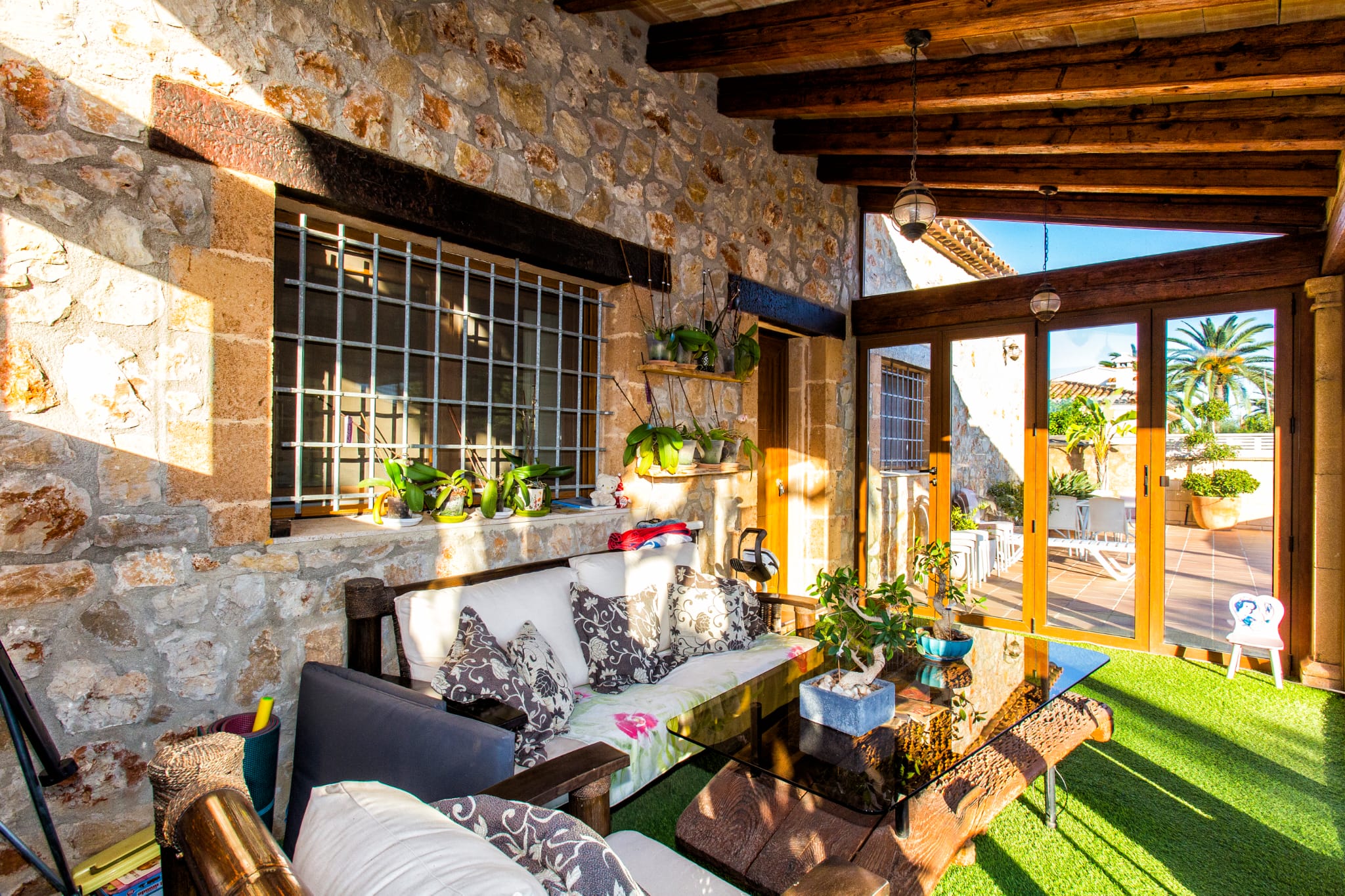 Covered terrace featuring exposed stone walls, dark wooden ceiling beams, and a seating area with a glass table on artificial turf, opening to a sunlit patio.