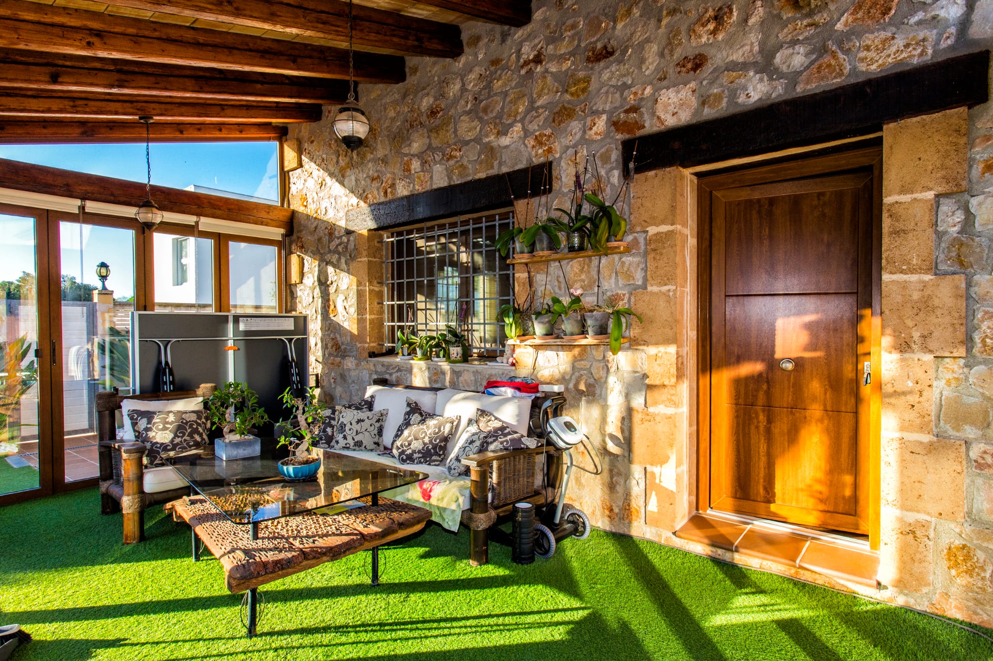 Covered terrace featuring exposed stone walls, wooden ceiling beams, and artificial turf. Includes a lounge area with a glass-topped driftwood table.