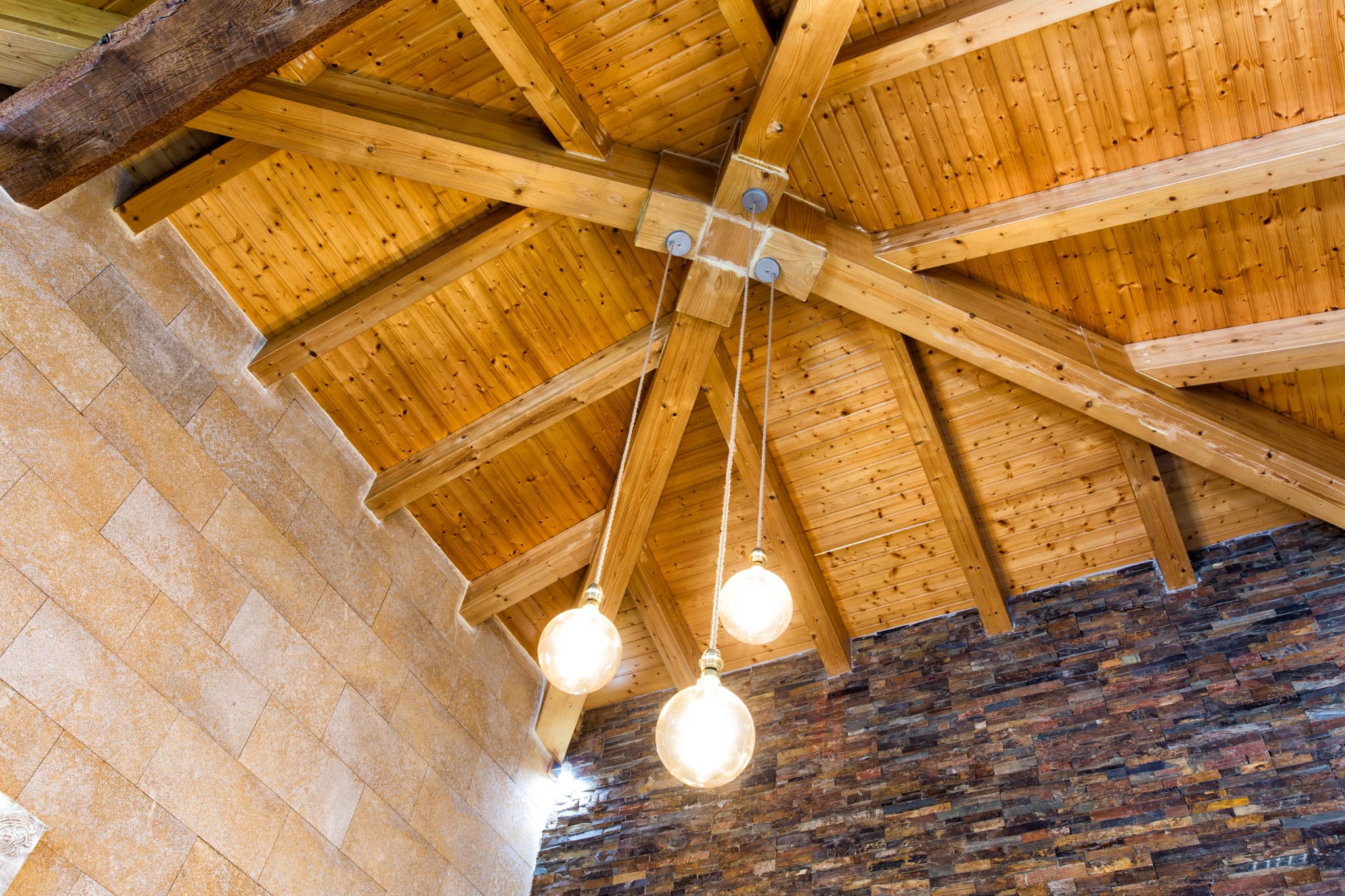 Detailed view of a vaulted ceiling featuring exposed pine beams in a radial layout, complemented by a trio of spherical pendant lights and textured stone walls.
