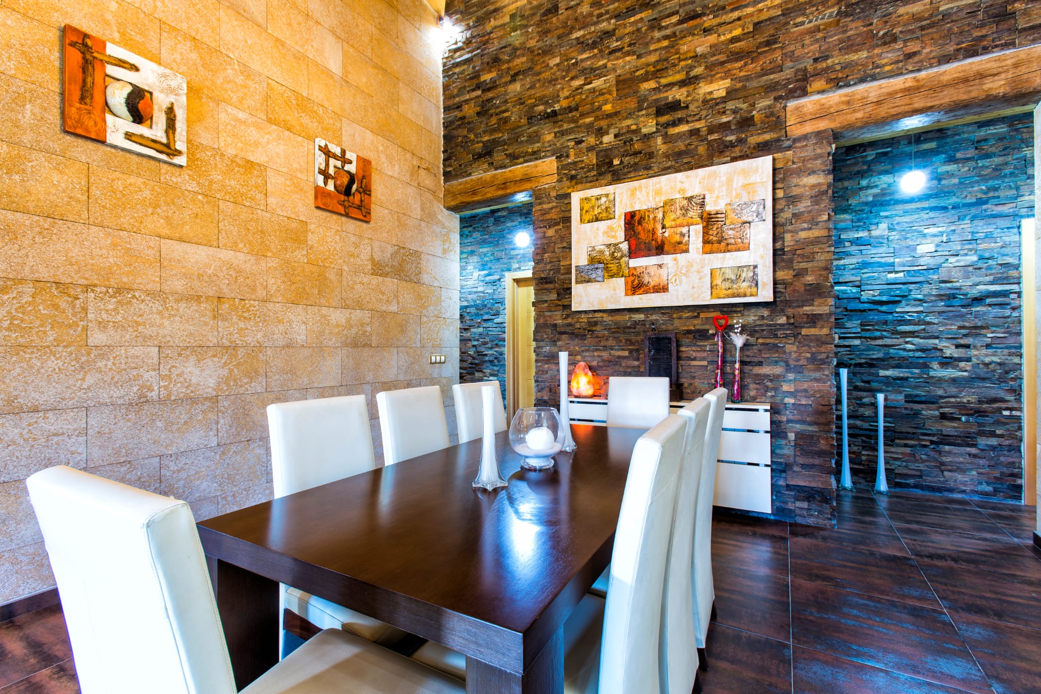 Dining room featuring a dark wood table with white chairs, set against textured stone and slate walls with abstract artwork.