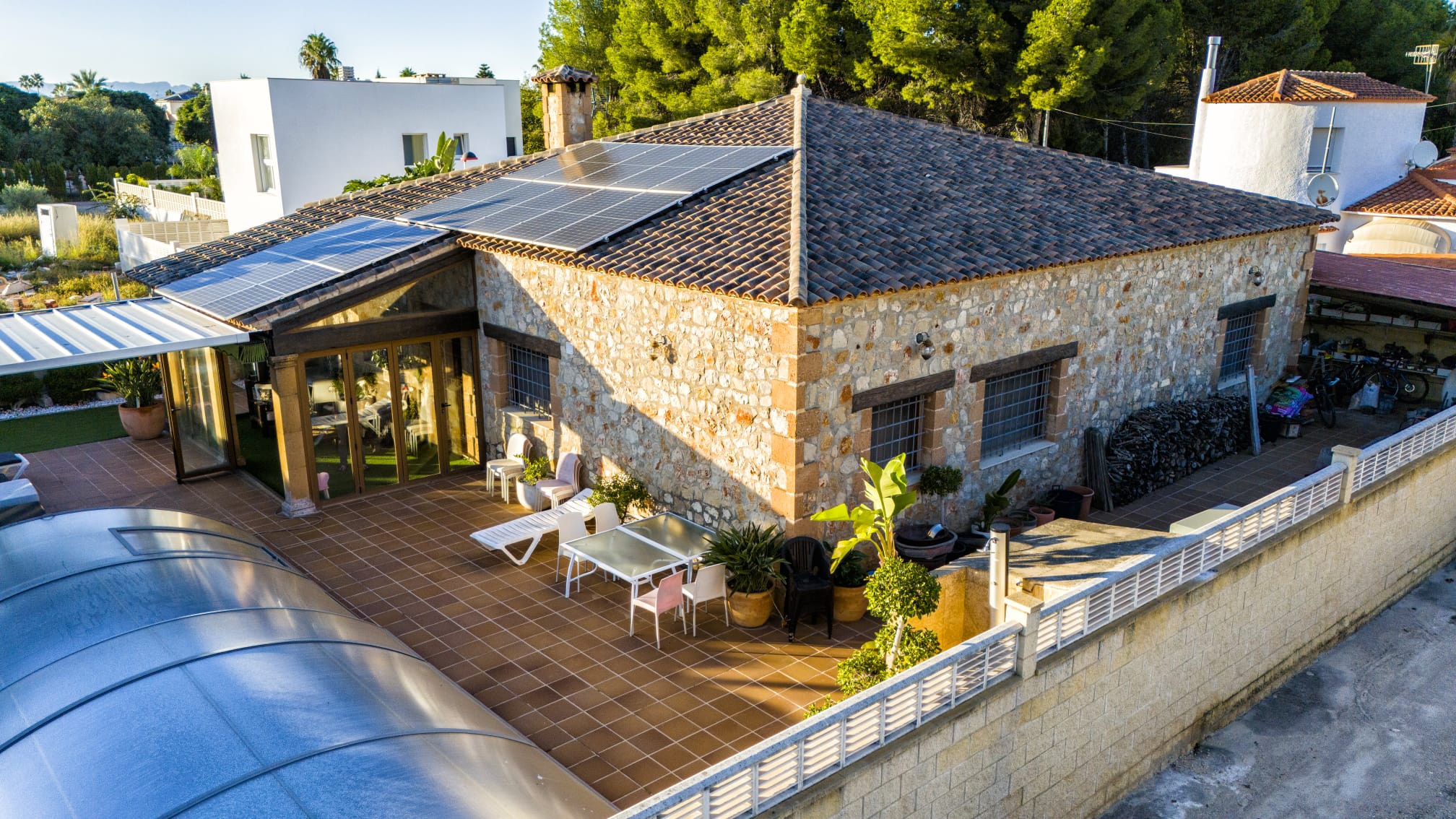 Exterior view of a stone villa featuring rooftop solar panels, a spacious tiled terrace, and a pool with a curved retractable cover.