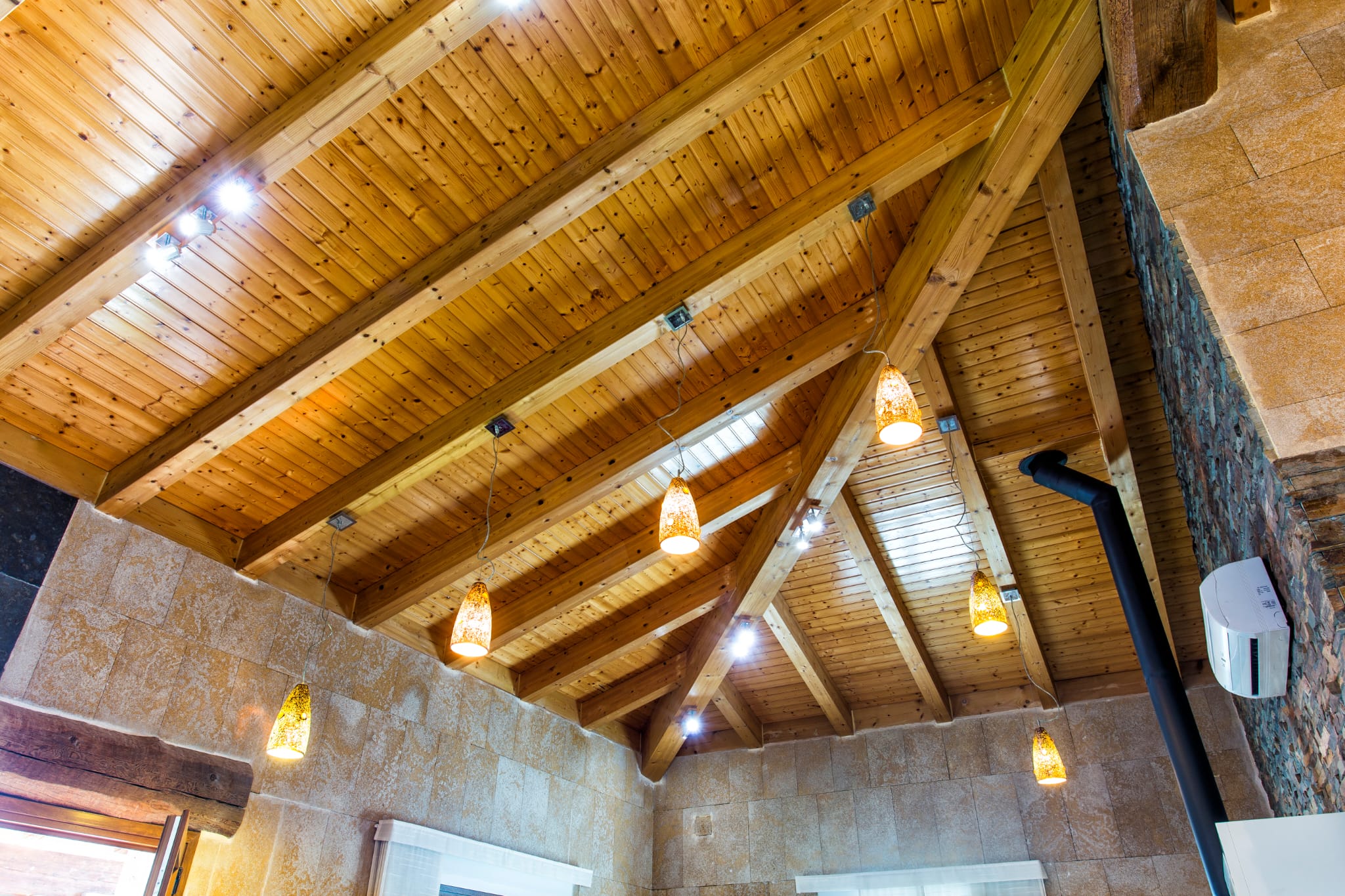 High vaulted ceiling featuring exposed pine beams and matching wood paneling, complemented by amber glass pendant lights and stone-clad walls.