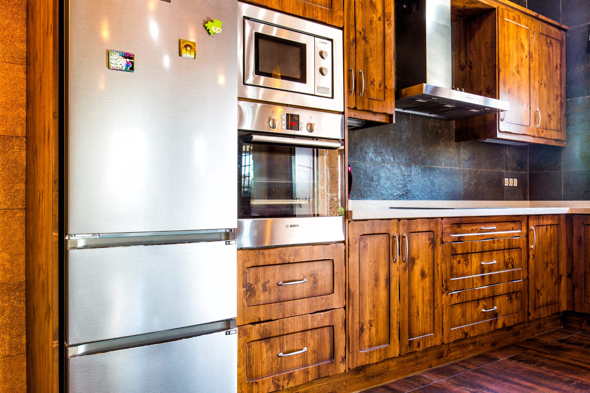 Kitchen featuring rustic wood cabinetry, integrated Bosch appliances in stainless steel, and a dark slate-textured backsplash with a matching range hood.