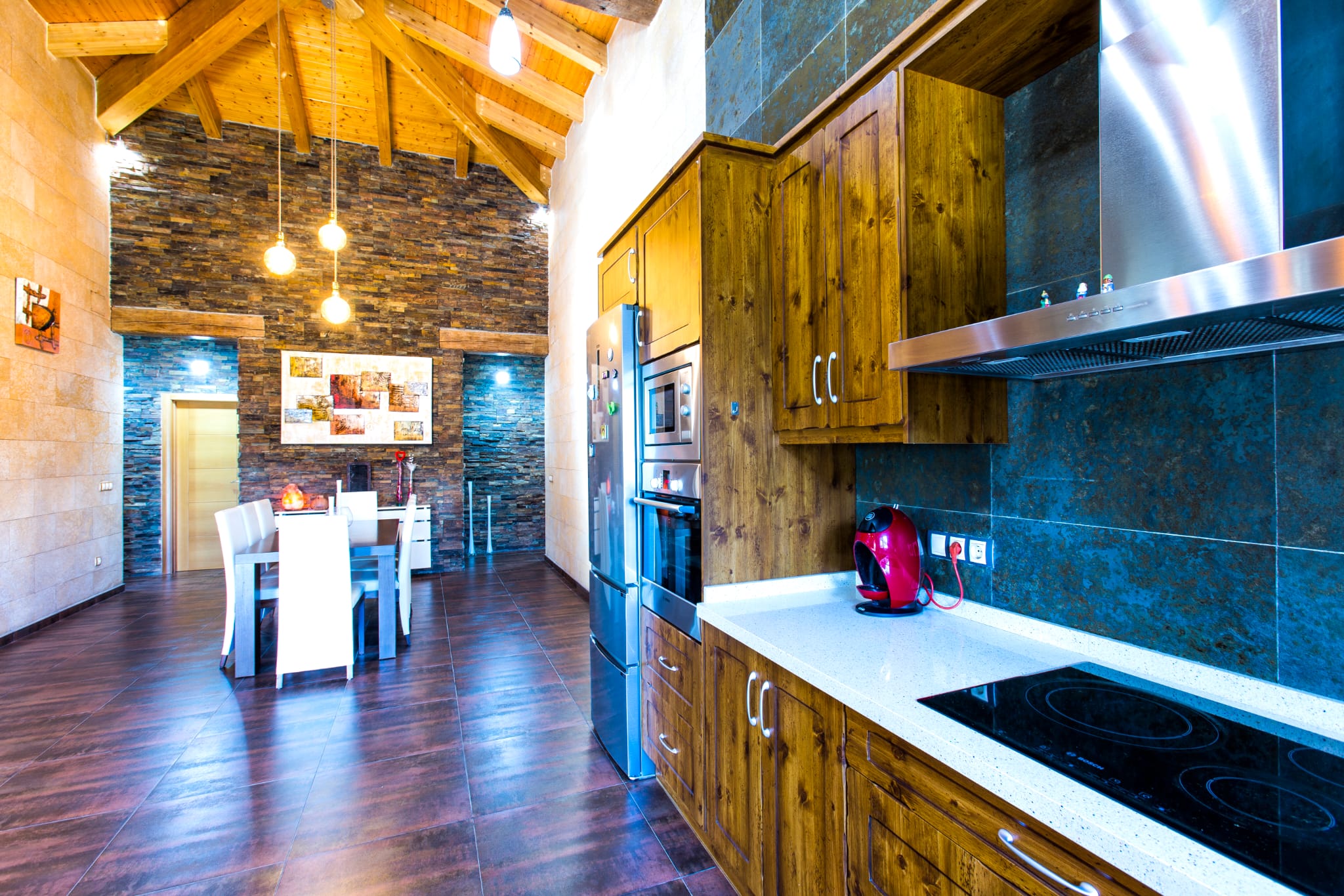 Kitchen featuring rustic wooden cabinets, a white countertop, and integrated stainless steel appliances, opening to a dining area with stone-clad walls and vaulted ceilings.