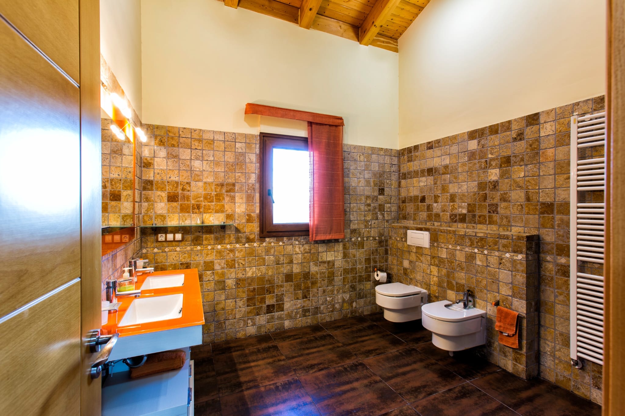 Modern bathroom featuring a double vanity with an orange countertop, stone-tiled walls, and a vaulted wooden ceiling with exposed beams.