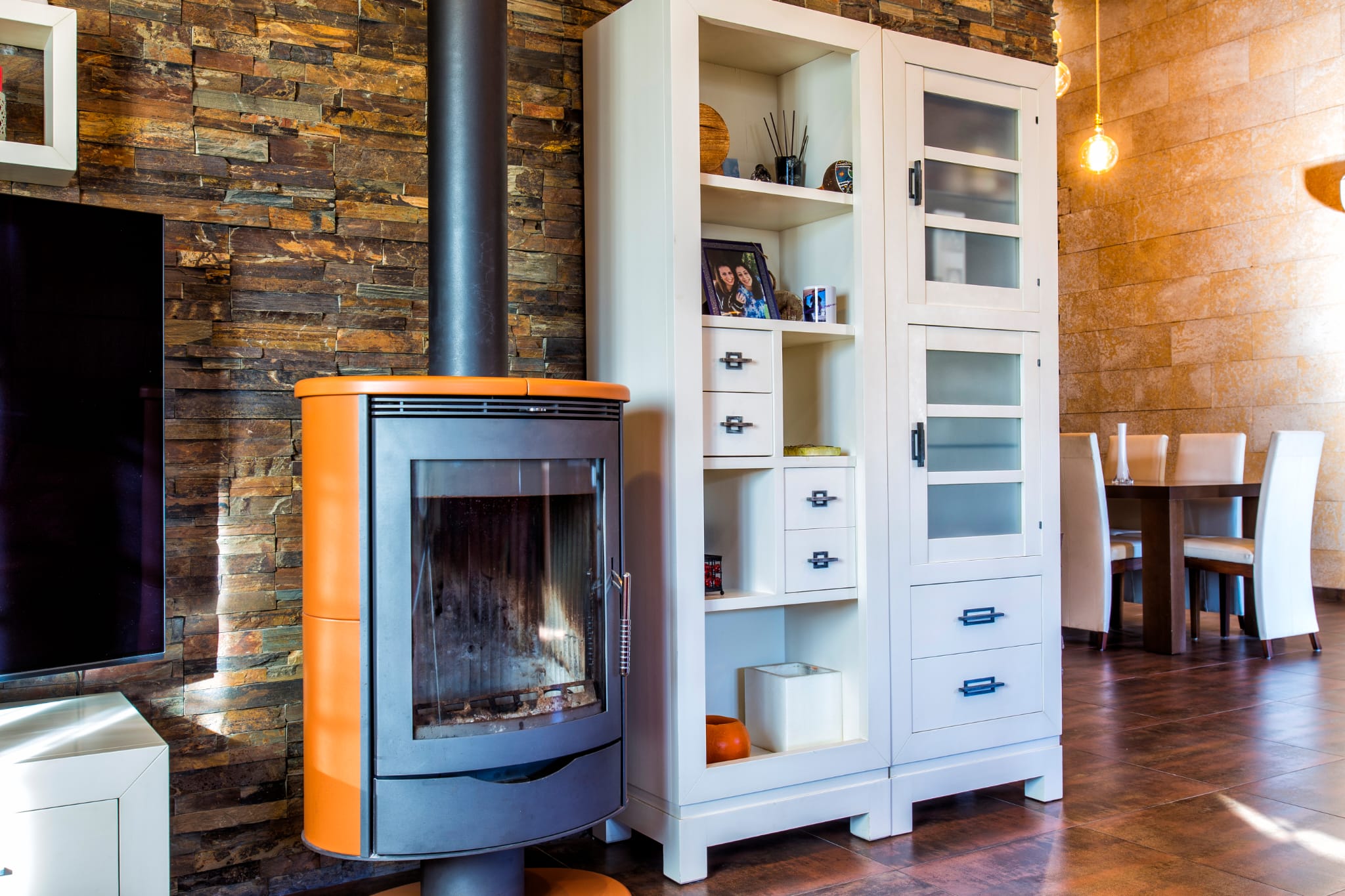 Modern wood-burning stove with orange accents set against a natural stone wall, adjacent to white cabinetry and a dining area with tiled flooring.