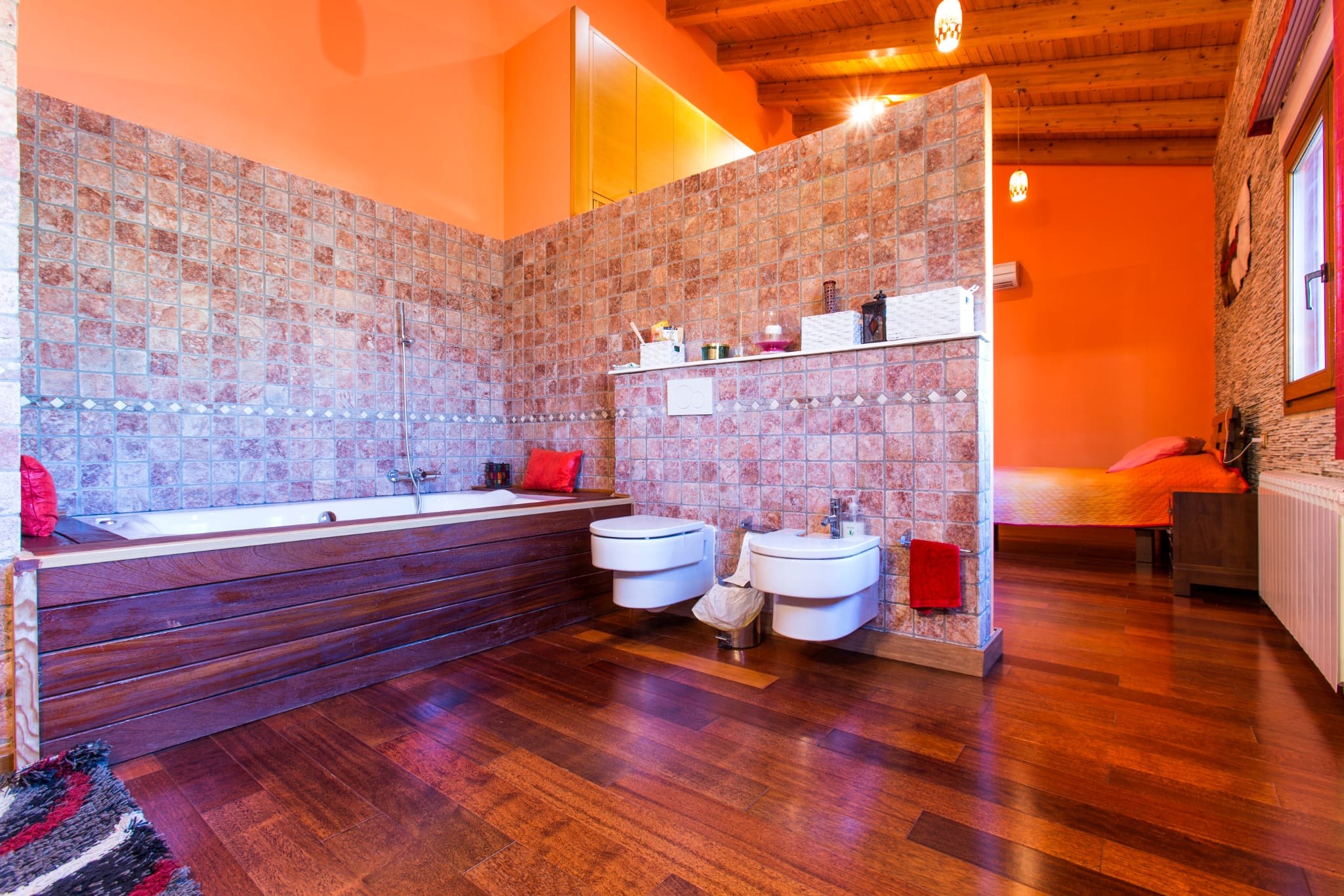 Open-concept bathroom featuring a wood-paneled bathtub, mosaic tile walls, and wall-hung fixtures, integrated with a bedroom under a beamed wooden ceiling.