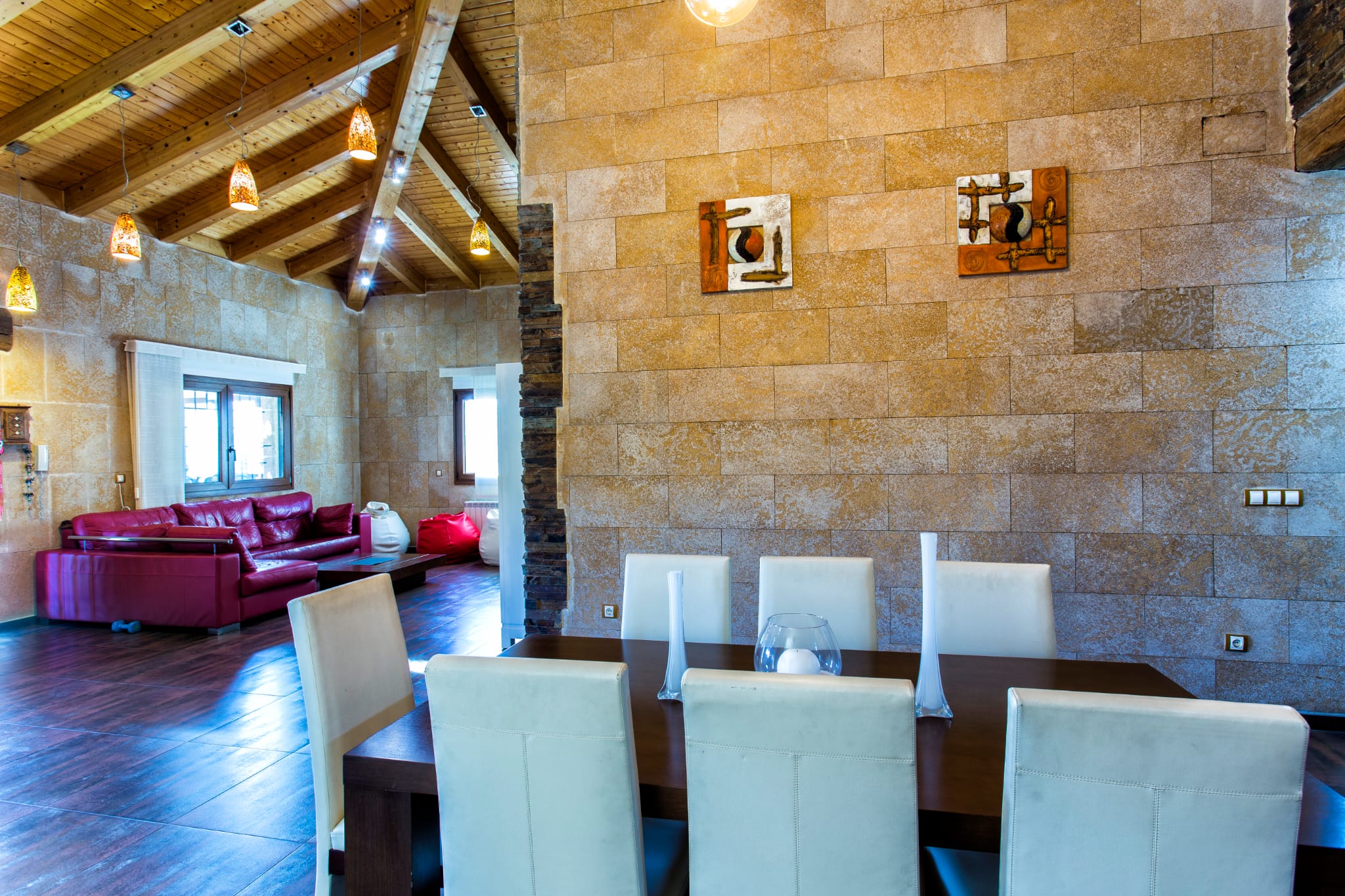 Open-plan dining area with textured stone walls, vaulted wooden ceilings with exposed beams, and a dark wood table paired with white high-back chairs.