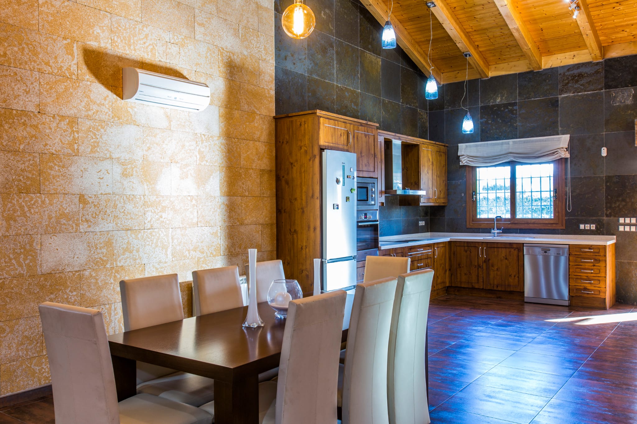 Open-plan kitchen and dining area featuring a vaulted wooden ceiling, stone-textured walls, rustic cabinetry, and modern stainless steel appliances.