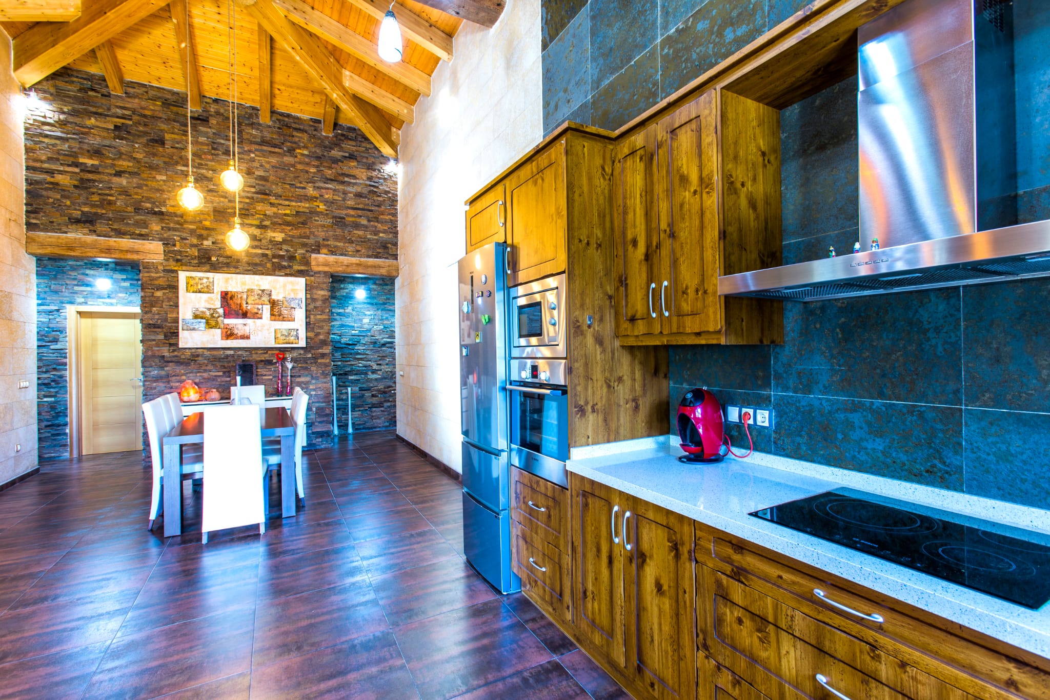 Open-plan kitchen featuring rustic wood cabinetry, integrated stainless steel appliances, and a vaulted beamed ceiling. A textured stone feature wall anchors the dining area.