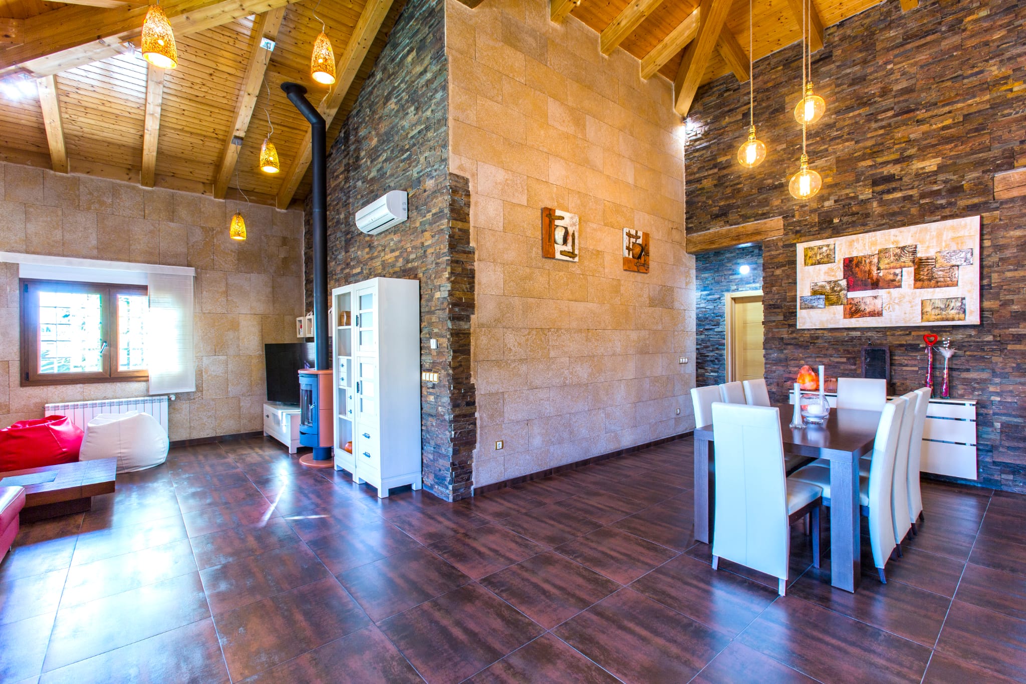 Open-plan living and dining area featuring vaulted wooden ceilings, natural stone walls, a modern wood stove, and warm pendant lighting over a dark wood dining table.