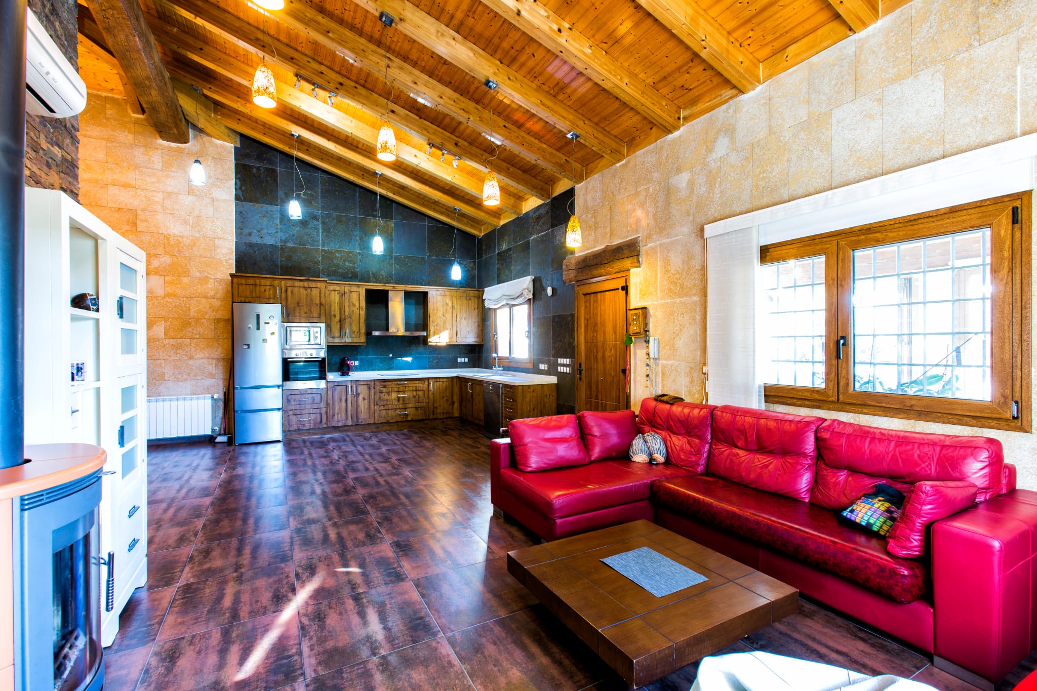 Open-plan living area featuring a vaulted wooden beam ceiling, red leather sofa, and a rustic kitchen with stone walls and integrated stainless steel appliances.