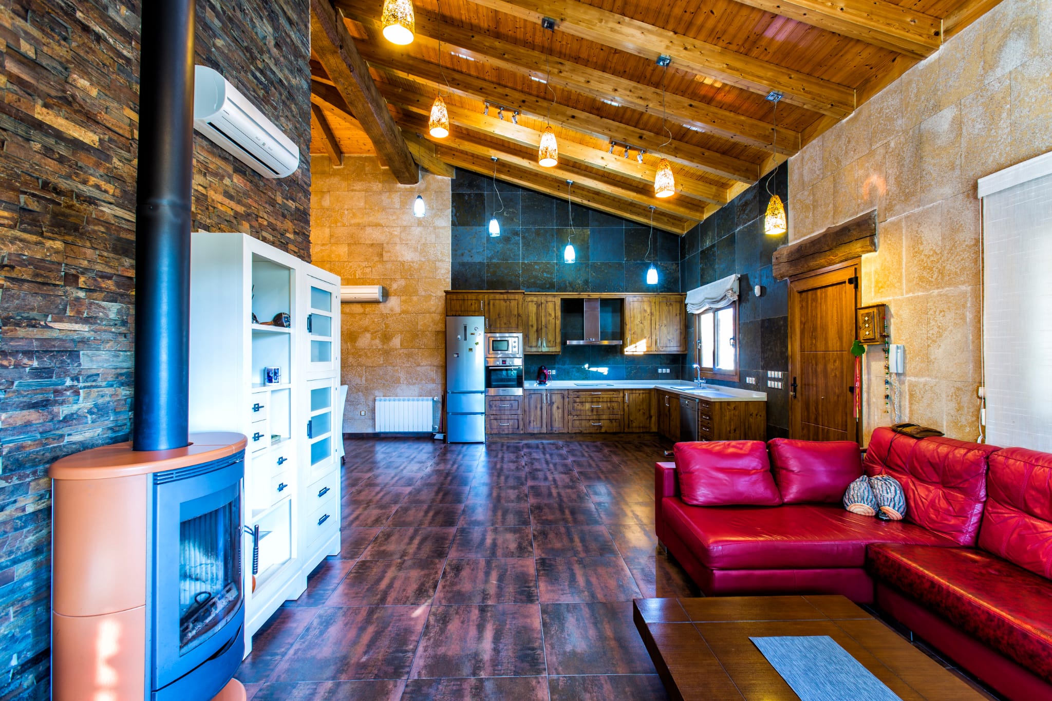 Open-plan living area featuring vaulted wooden ceilings, a modern wood-burning stove, and a rustic kitchen with integrated appliances and natural stone wall cladding.