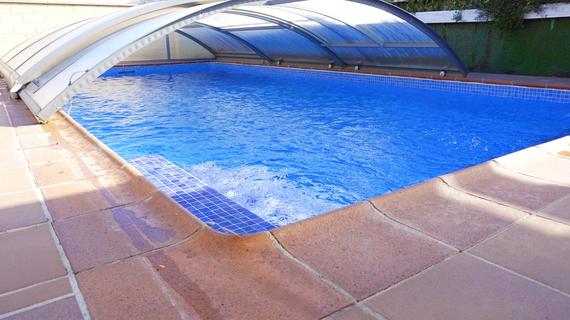 Outdoor swimming pool featuring a retractable telescopic enclosure, blue mosaic tile interior, and a terracotta stone deck.