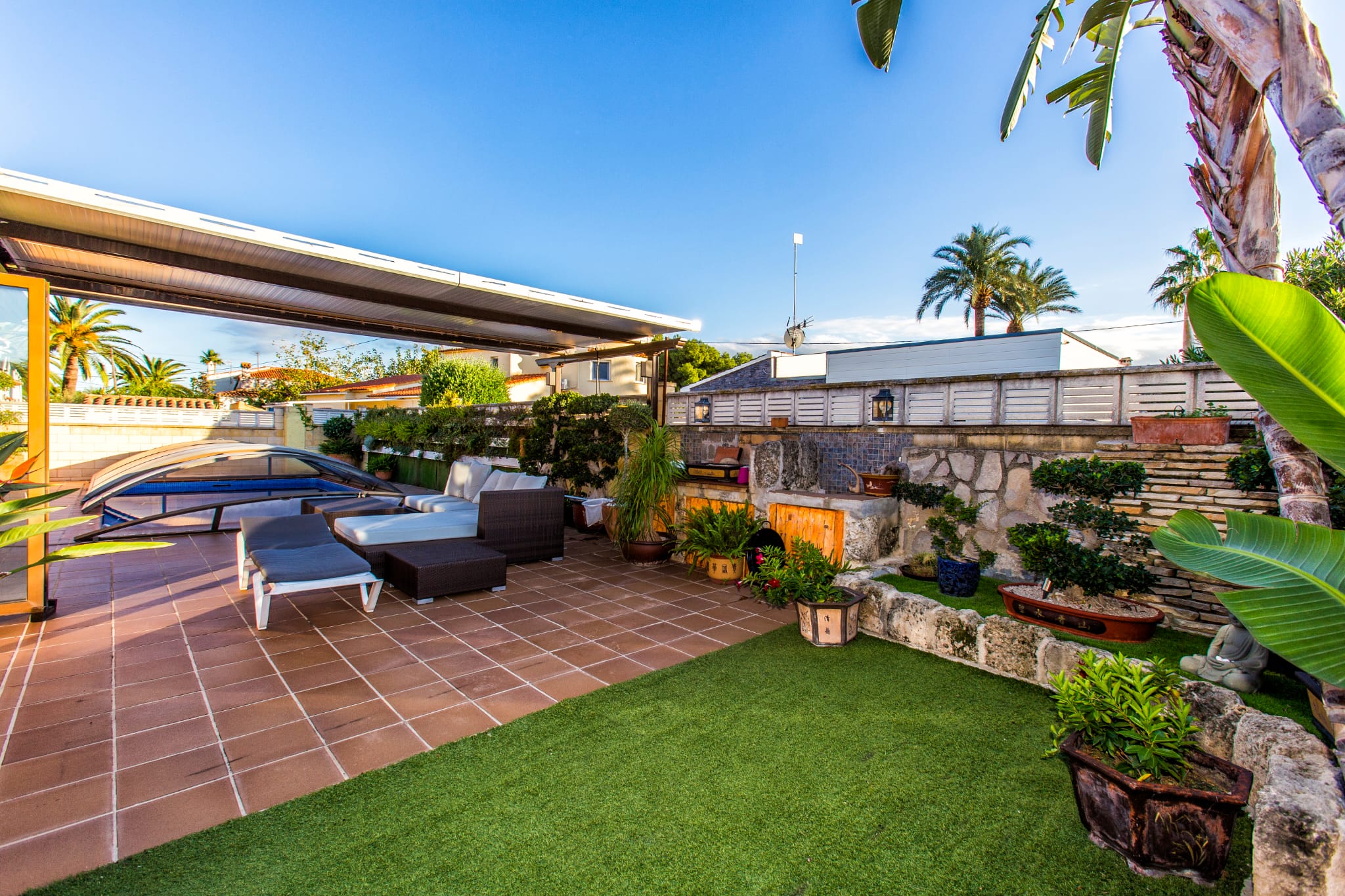 Outdoor terrace featuring terracotta tiling, artificial turf, and a lounge area under a pergola, adjacent to a pool with a retractable cover and stone planters.