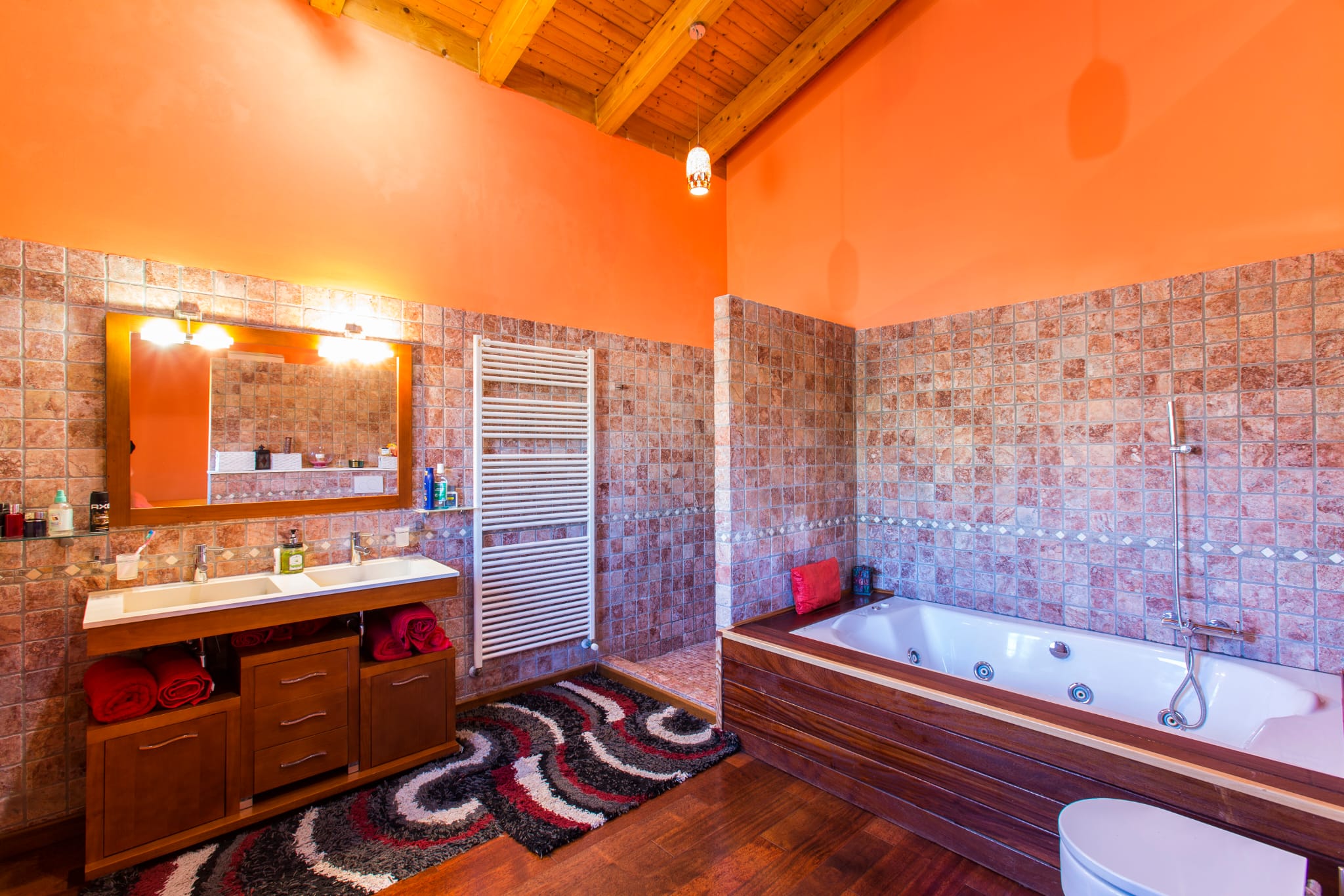 Rustic bathroom featuring exposed wooden ceiling beams, a double vanity with dark wood cabinetry, a whirlpool bathtub, and textured stone-effect wall tiling.