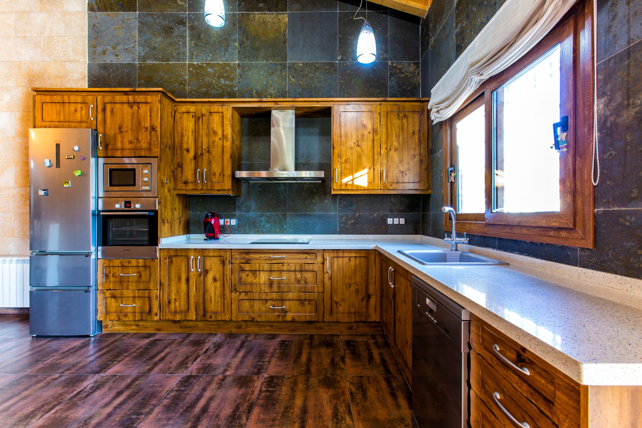 Rustic kitchen featuring knotty wood cabinetry, dark stone wall tiles, and integrated stainless steel appliances under a vaulted ceiling with pendant lighting.