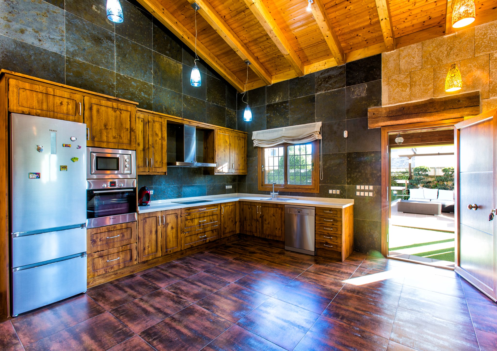 Rustic kitchen featuring wooden cabinetry, dark slate wall tiles, and a beamed ceiling. Includes modern stainless steel appliances and direct access to an outdoor patio.