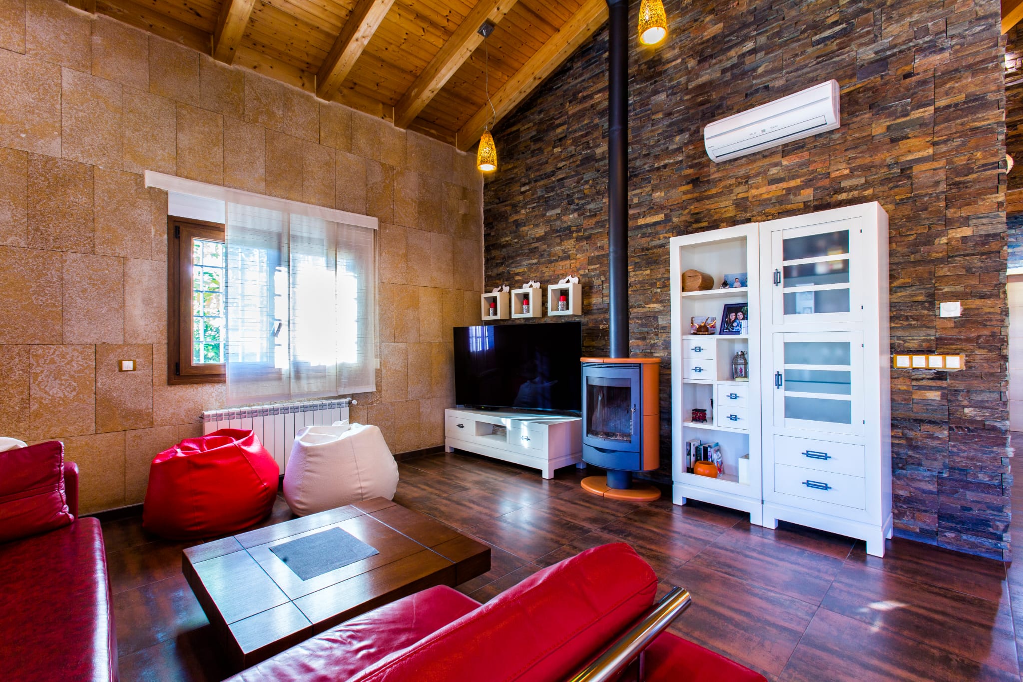 Rustic living room featuring natural stone walls, exposed wooden ceiling beams, a modern wood-burning stove, and vibrant red leather seating.