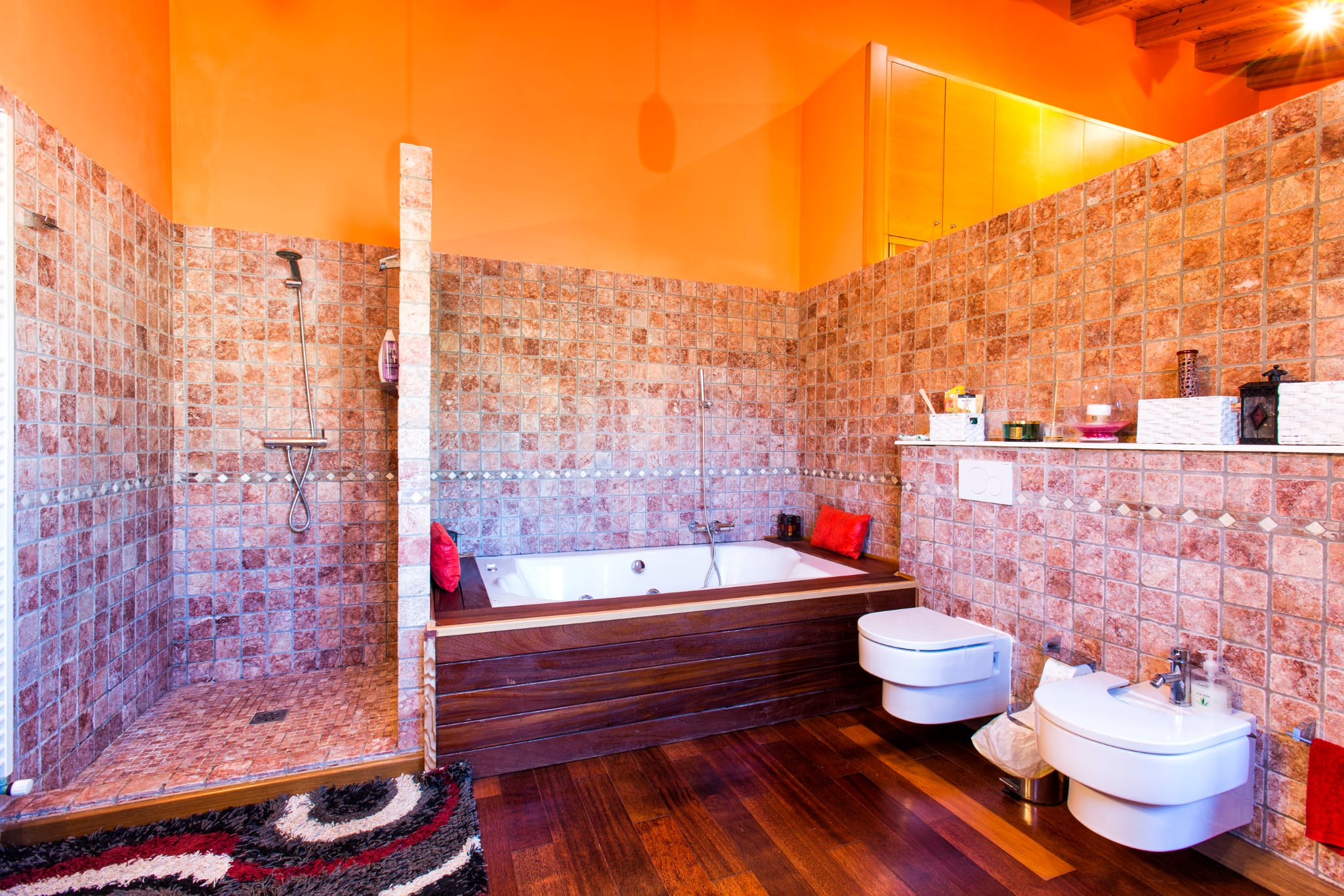 Spacious bathroom featuring a walk-in stone tile shower, a bathtub with dark wood paneling, and wall-mounted fixtures against vibrant orange walls and wood flooring.