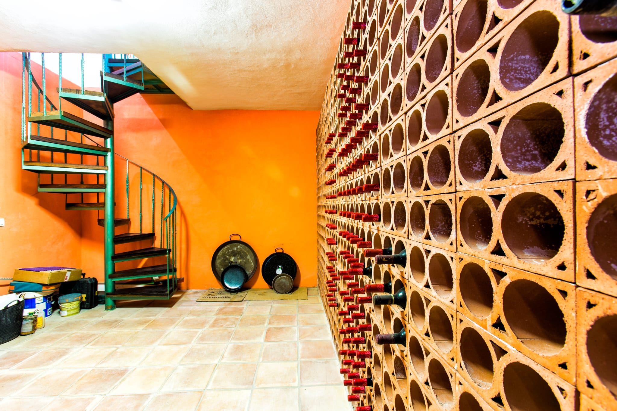 Wine cellar featuring a terracotta honeycomb bottle rack, an orange accent wall, and a green metal spiral staircase with dark wooden steps.