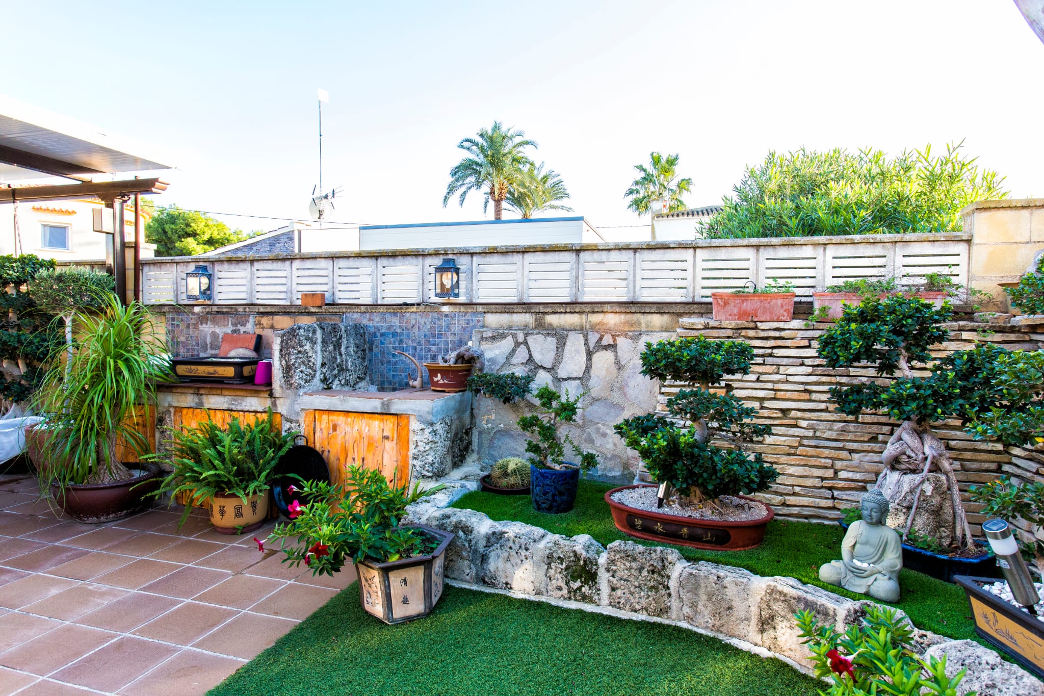 Zen-style patio garden featuring artificial turf, stacked stone walls, and a collection of bonsai trees. Includes a built-in stone counter with blue mosaic tiles.