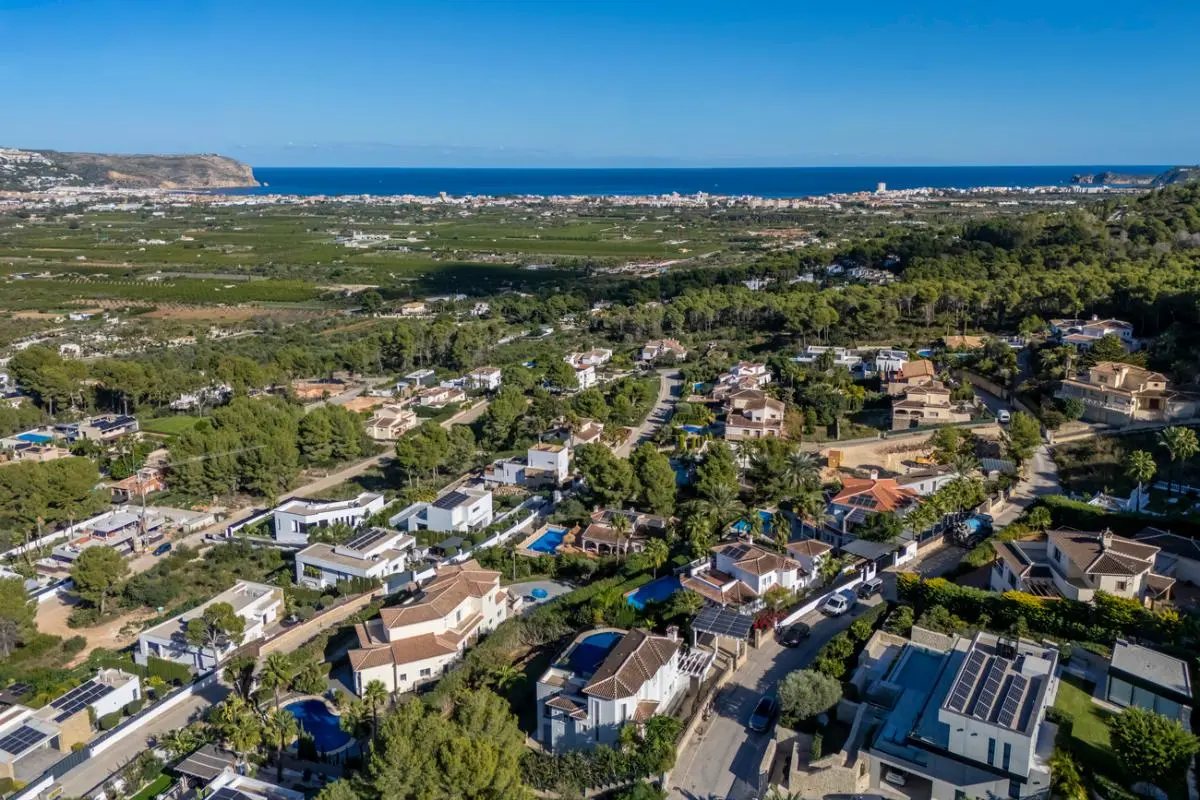 Aerial perspective of a hillside residential neighborhood featuring villas with solar panels and private pools, overlooking a coastal valley and the sea.
