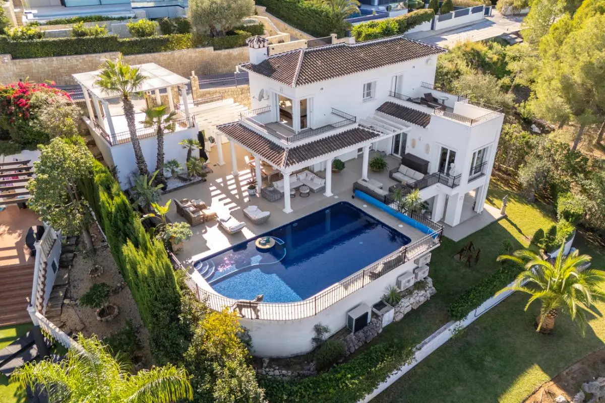 Aerial view of a white Mediterranean villa featuring a large swimming pool, multiple covered terraces with columns, and landscaped gardens with palm trees.