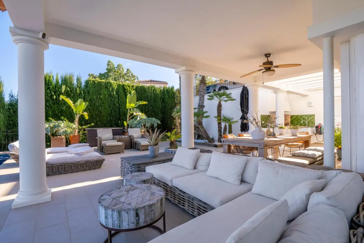 Covered terrace featuring white columns, a large wicker sectional sofa, and an outdoor dining area with a ceiling fan, surrounded by tropical greenery.