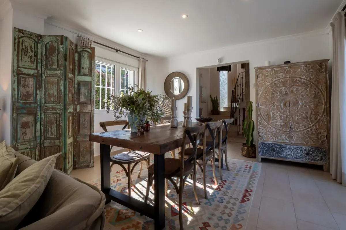 Dining area featuring a rustic wooden table, cross-back chairs, and a large hand-carved cabinet. A vintage green screen and patterned rug complement the neutral tiled flooring.
