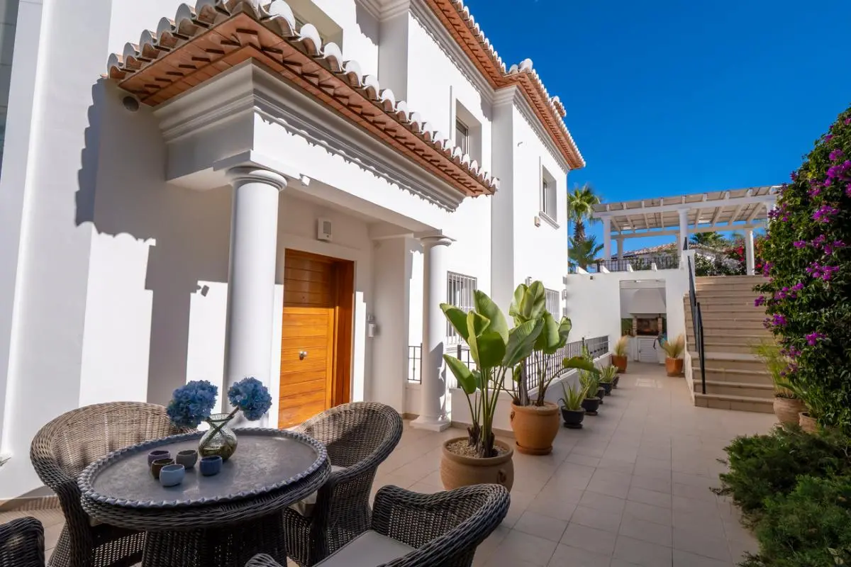 Exterior patio featuring a wicker seating area, white architectural columns, and a wooden entrance door. Potted plants line the walkway leading to a pergola.