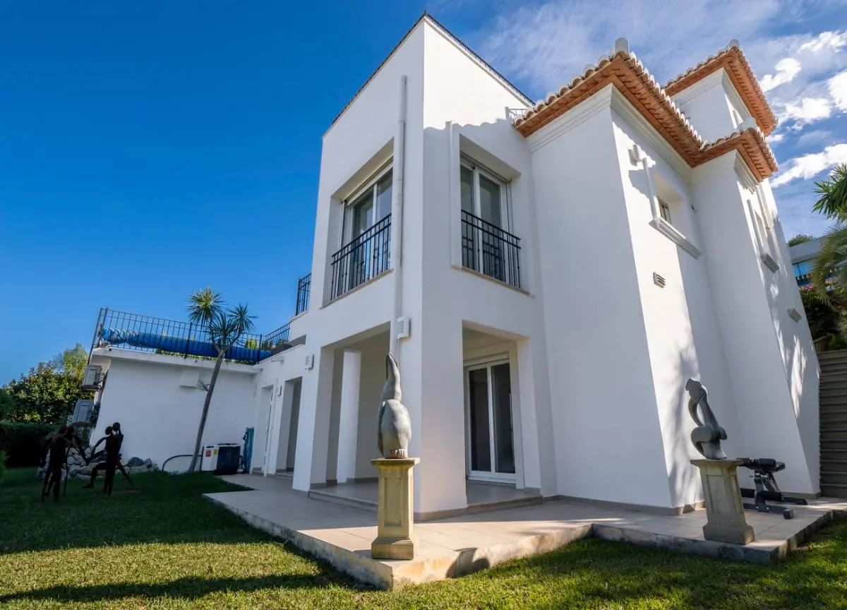 Exterior view of a multi-level white villa featuring terracotta roof tiles, black iron railings, and a manicured lawn with decorative stone sculptures.