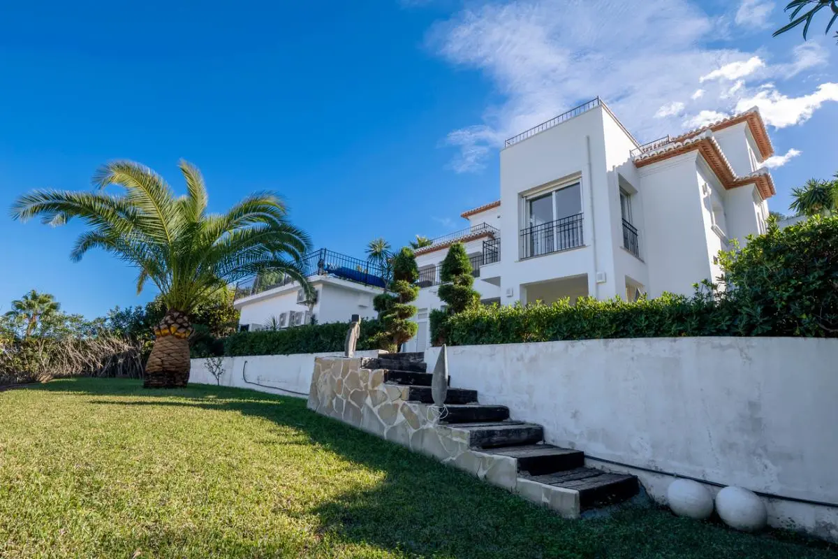 Exterior view of a multi-level white villa featuring terracotta roof tiles, black metal railings, and a manicured lawn with a mature palm tree.