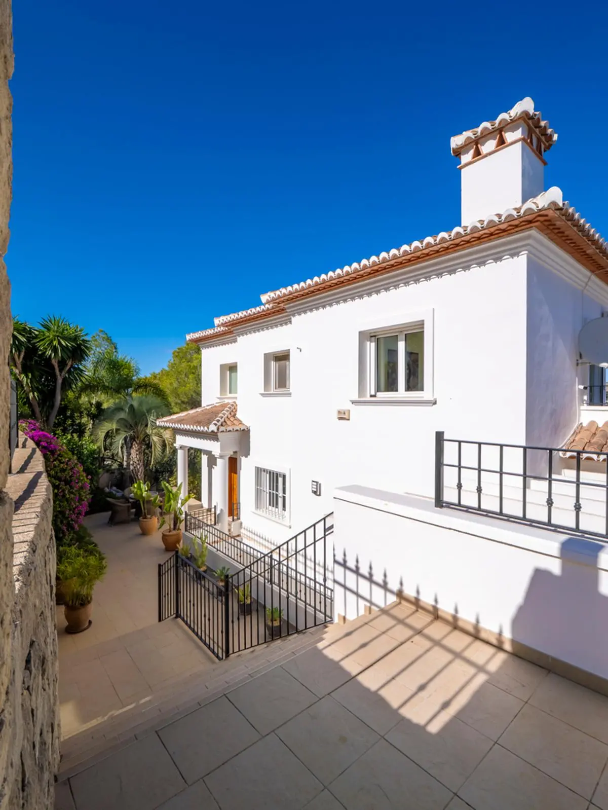 Exterior view of a white Mediterranean villa featuring terracotta roof tiles, a decorative chimney, and black wrought iron railings overlooking a tiled terrace.
