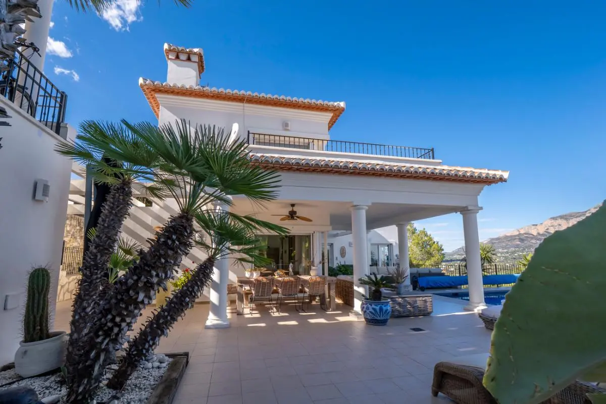 Exterior view of a white villa featuring a covered terrace with columns, an outdoor dining area, and Mediterranean landscaping overlooking the mountains.