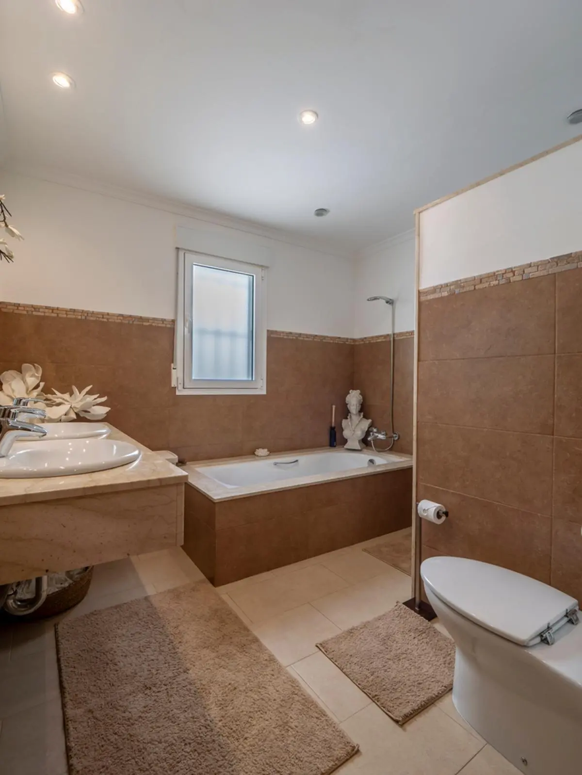Full bathroom featuring a marble double vanity, integrated bathtub with brown tiling, recessed lighting, and a frosted privacy window.