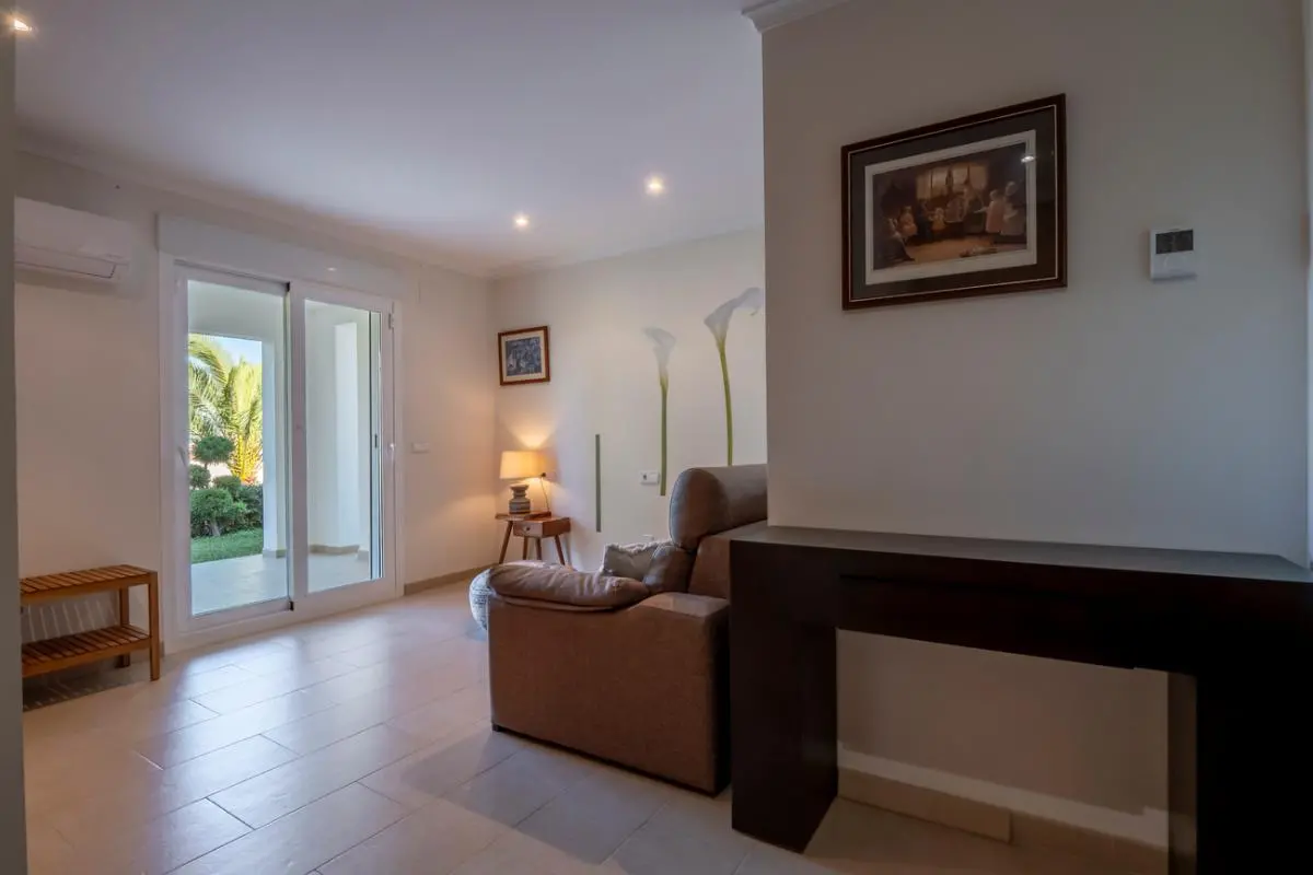 Living area featuring a dark wood console, brown leather seating, and tiled floors. A sliding glass door provides direct access to the garden and natural light.