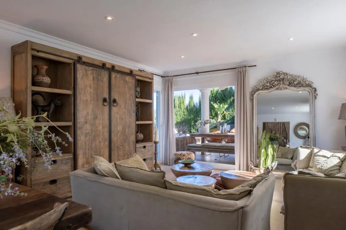 Living area featuring a rustic wooden cabinet with sliding doors, neutral sofas, and an ornate floor mirror, opening onto a terrace with outdoor dining.